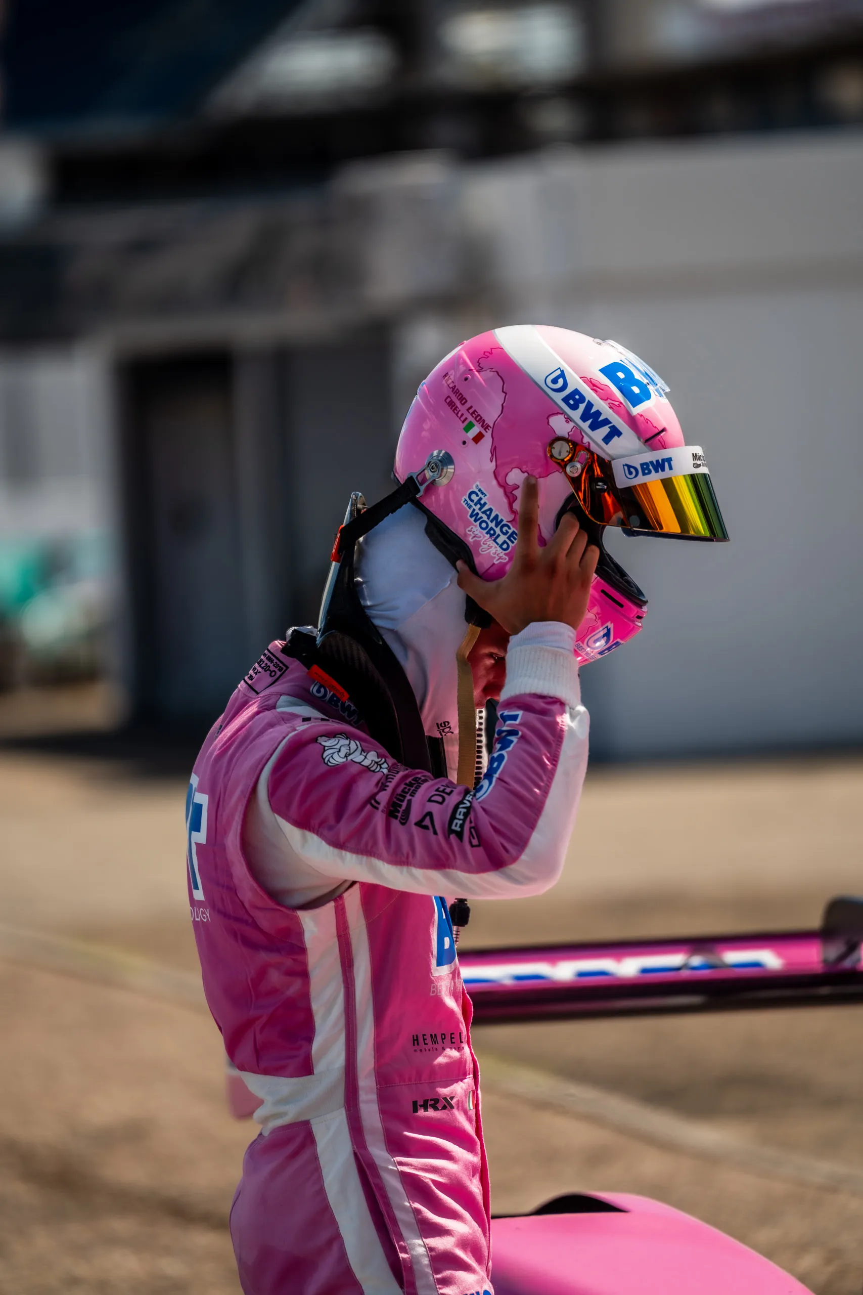 Riccardo Leone Cirelli, wearing a pink-and-white racing suit, is shown in profile reaching with both hands for his helmet with a mirrored visor; behind him is the rear wing of his pink race car.