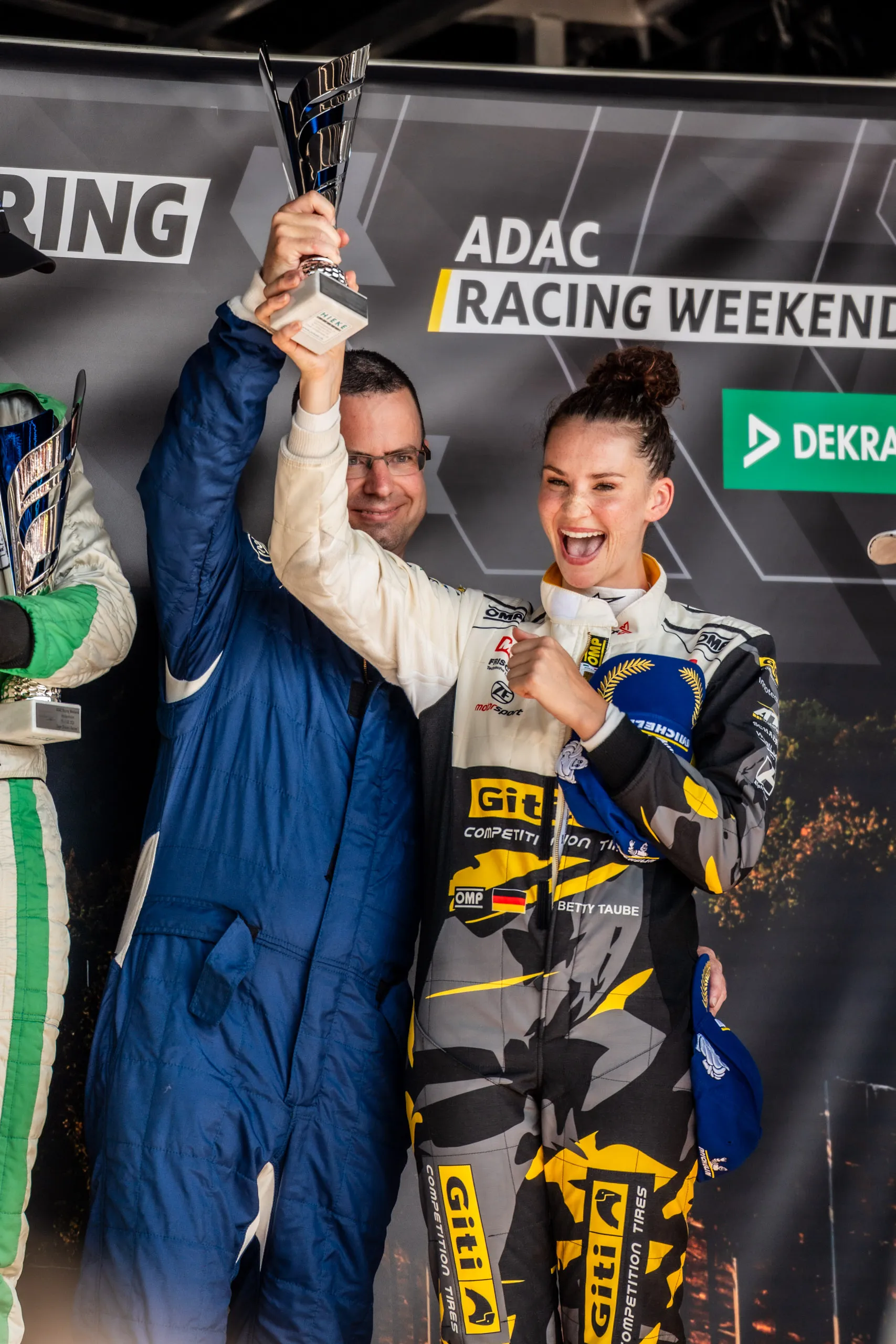 Betty Taube, wearing a gray-and-yellow racing suit, celebrates on the podium; next to her, a man in a blue racing suit holds a trophy aloft. In the background is the ADAC Racing Weekend banner