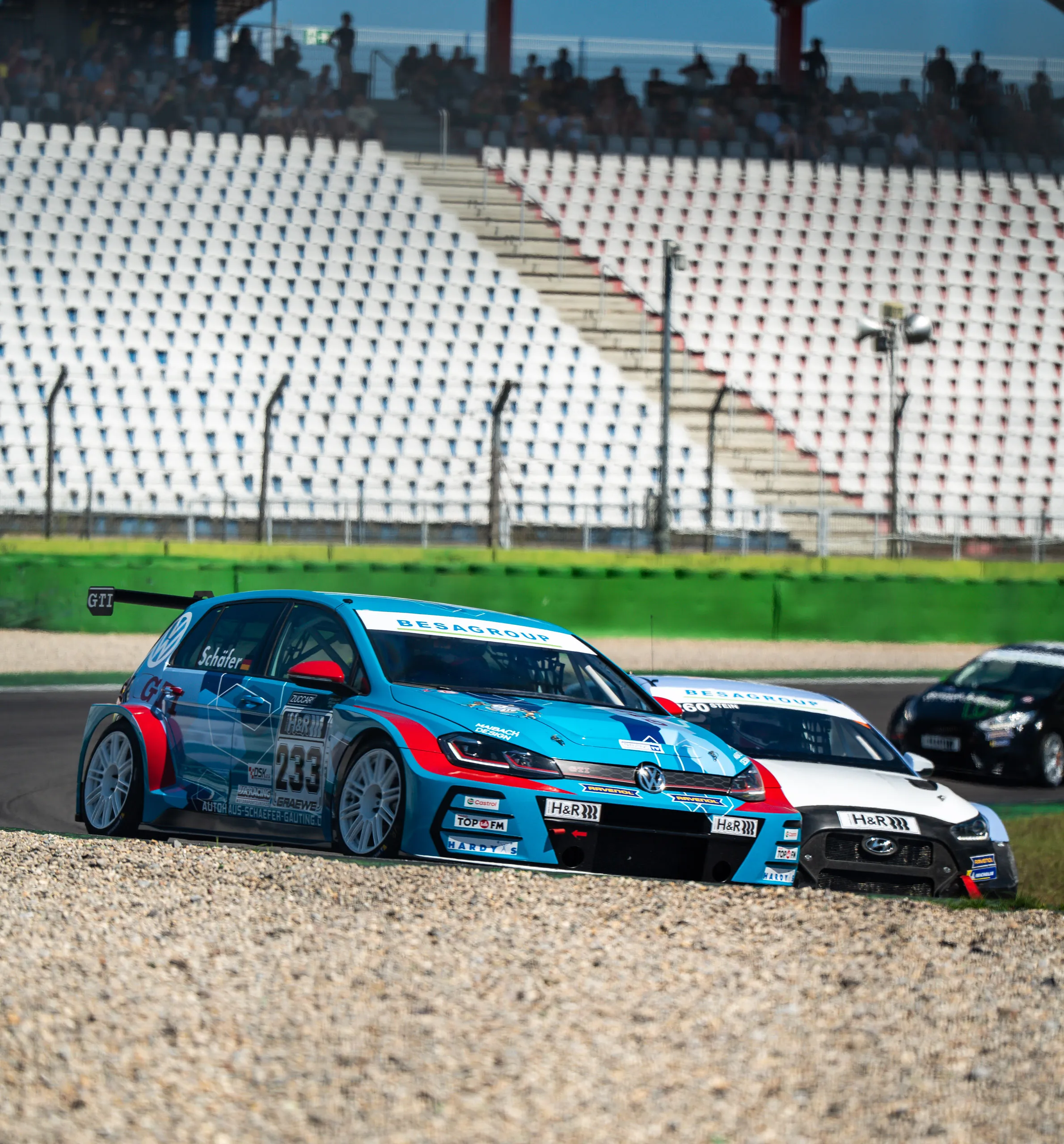 A blue VW Golf GTI TCR with start number 233 is racing through a turn right in front of a white Hyundai, with the largely empty grandstands of the Hockenheimring in the background.