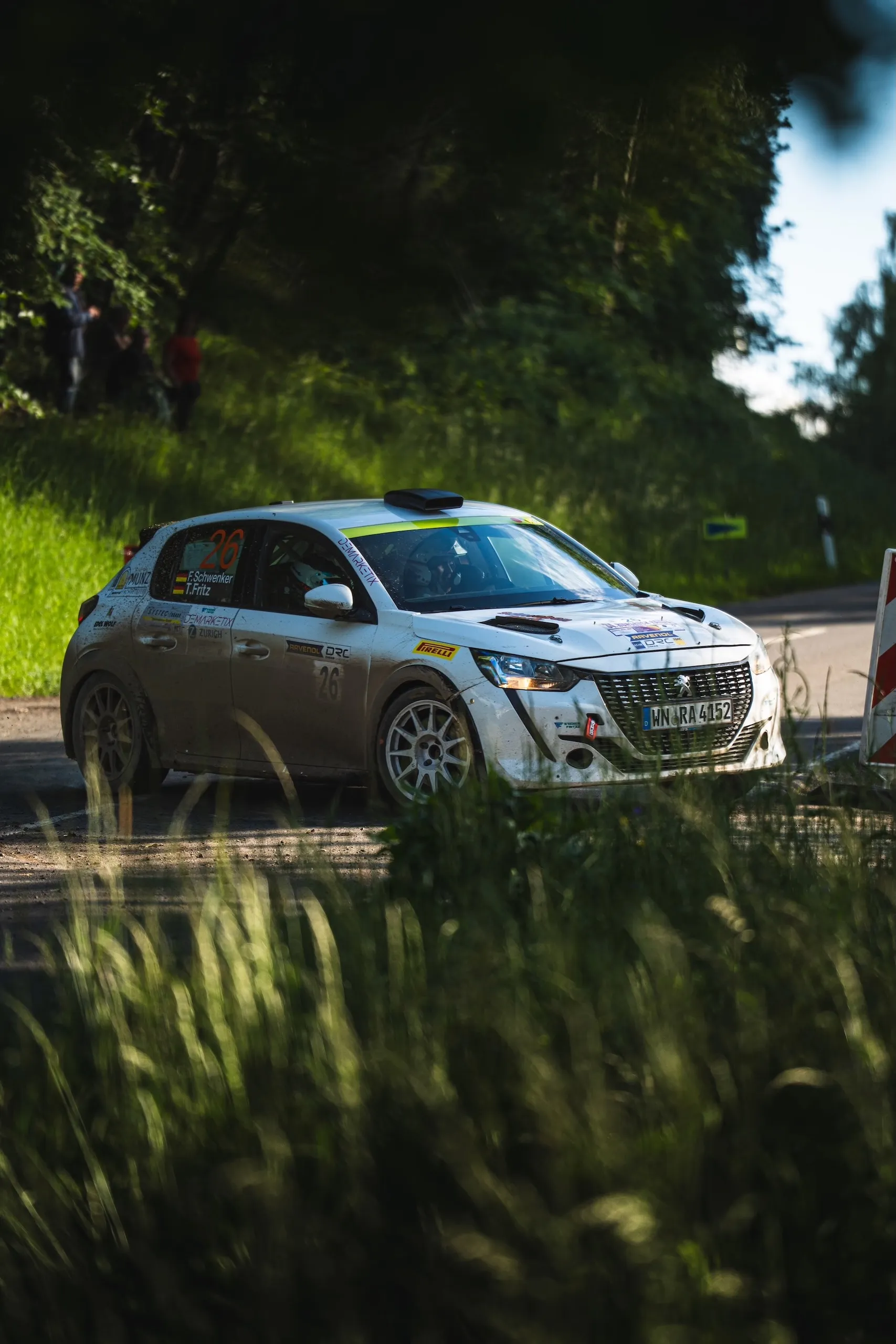 A mud-covered Peugeot 208 Rally4 with start number 26, driven by Florian Schwenker and Tobias Fritz, in the evening light of a wooded special stage.