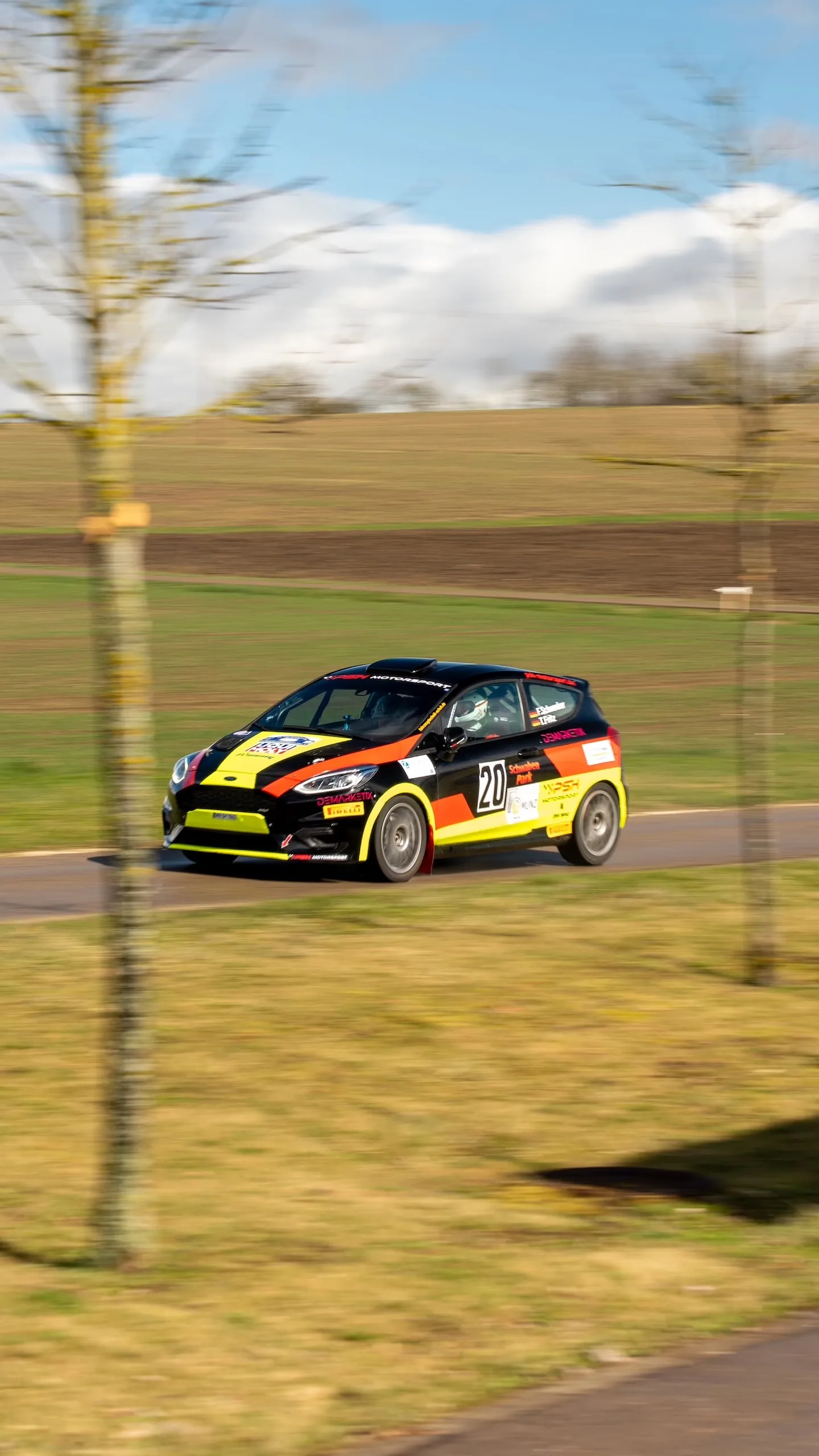 A Ford Fiesta Rally4 with start number 20, driven by Florian Schwenker and Tobias Fritz, speeding down a country road, with fields stretching out in the background under a blue sky.