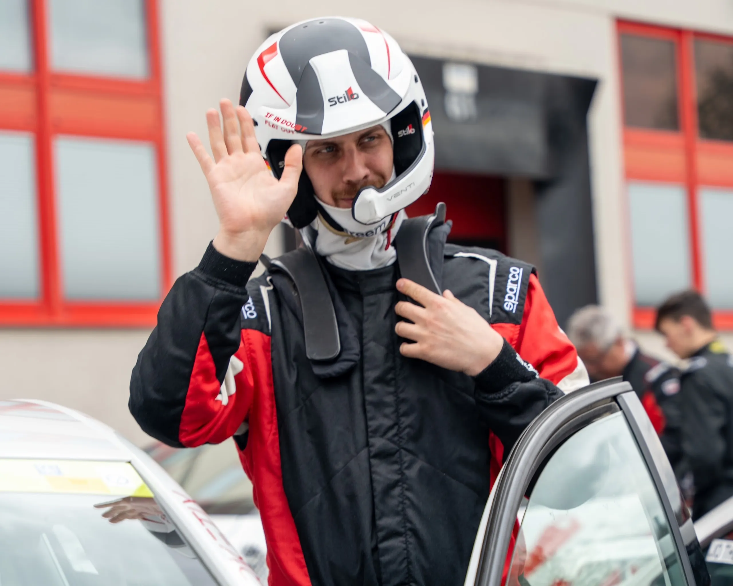 Florian Schwenker in his racing suit with his helmet on, just before getting into the car, raising his hand in greeting.