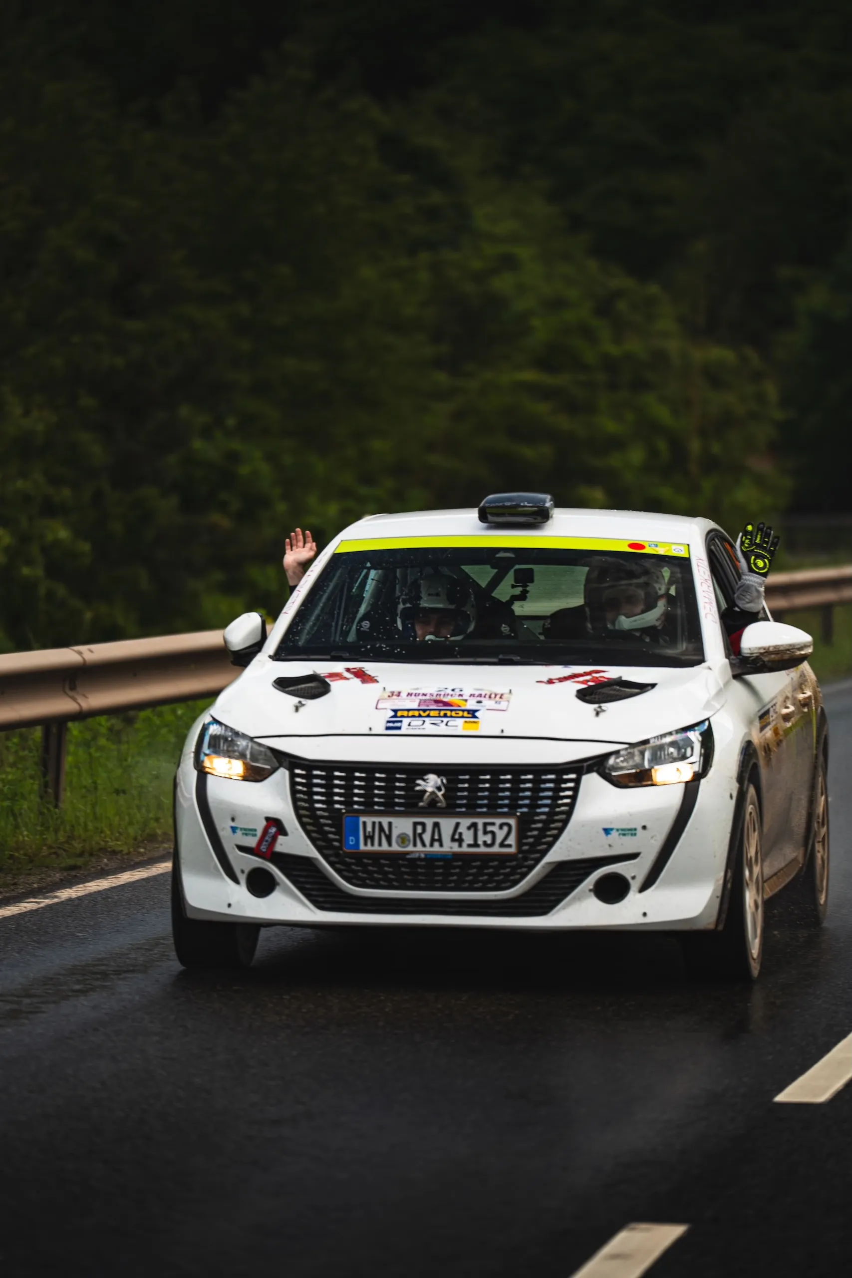 Florian Schwenker and Tobias Fritz’s Peugeot 208 Rally4 on a rain-soaked road during the 34th Hunsrück Rally; both are waving out the windows.