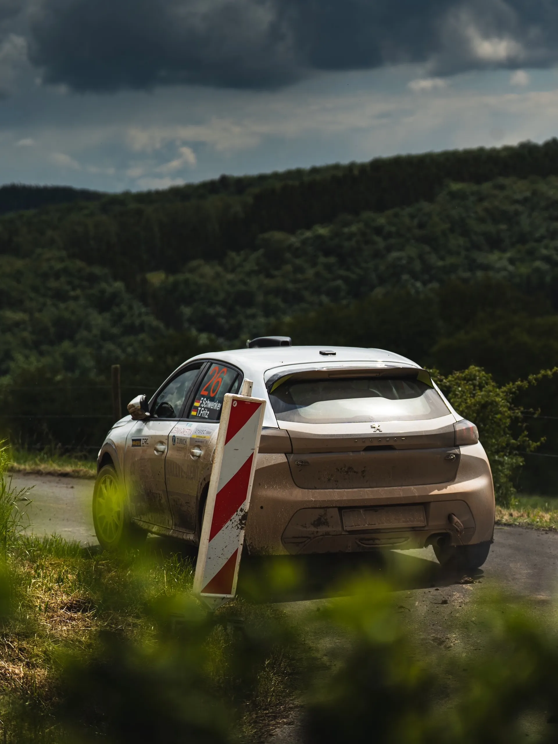 Rear view of Florian Schwenker and Tobias Fritz’s mud-covered Peugeot 208 Rally4 in a curve, with forested hills behind it under dark clouds.