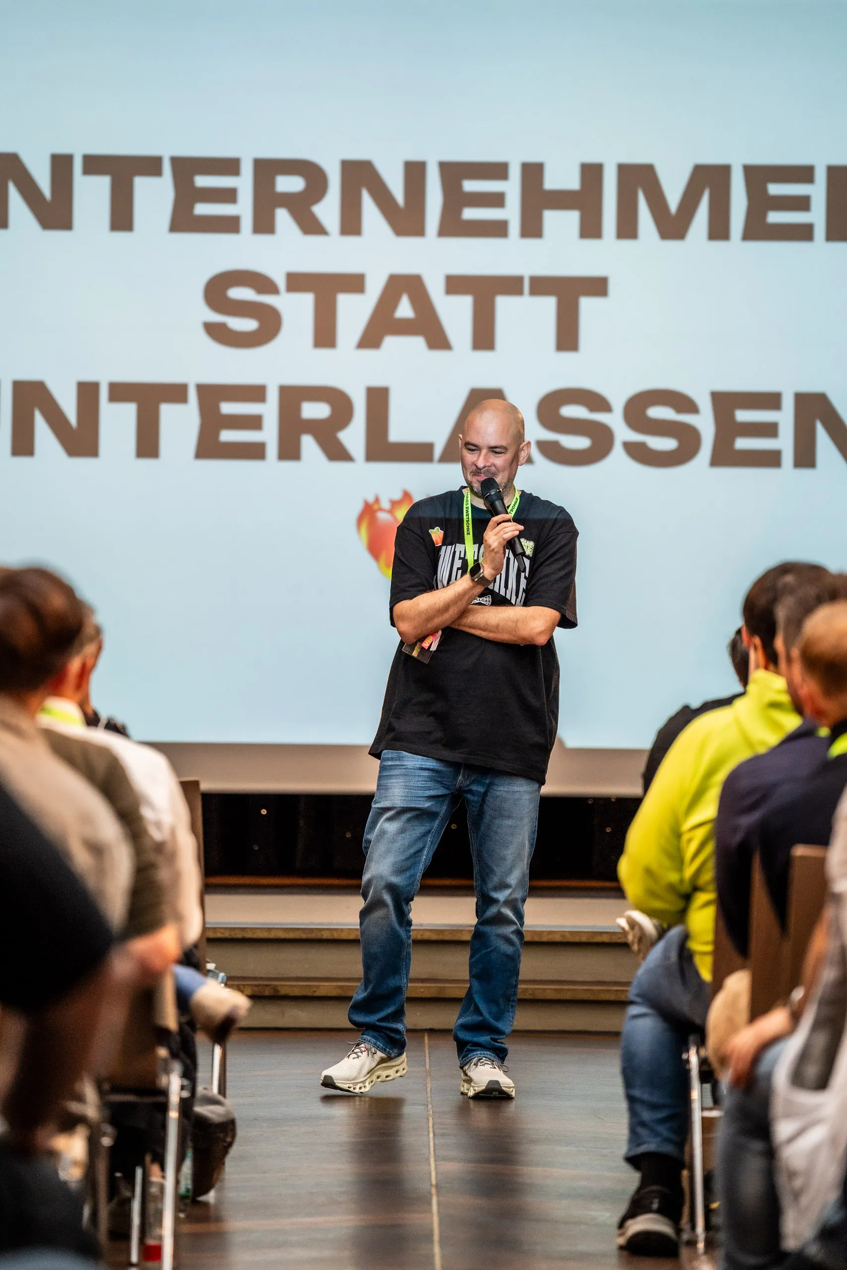 Hannes Zwetschke steht mit Mikrofon vor dem Publikum des Community-Day 2025, im Rücken die Leinwand mit dem Schriftzug „Unternehmen statt Unterlassen".
