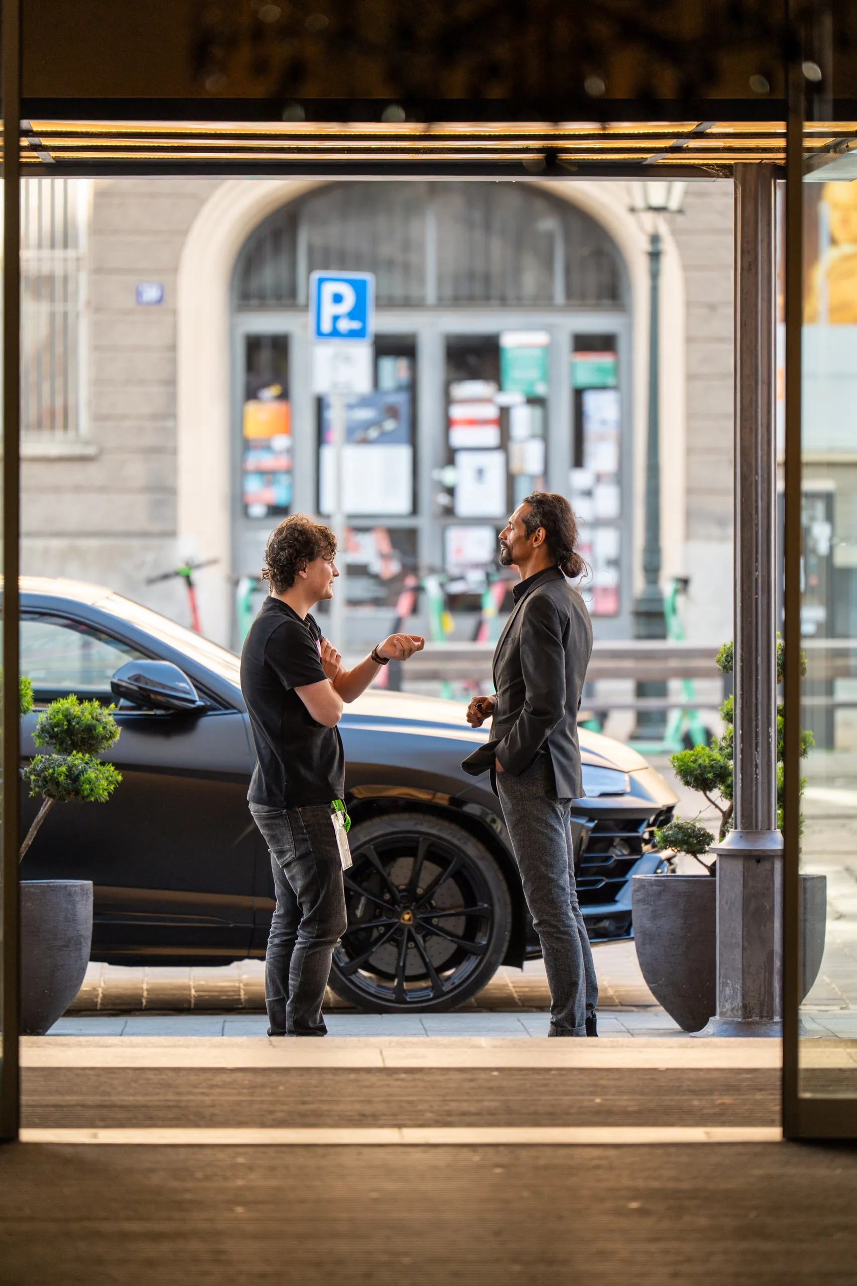 Zwei Teilnehmer des Hannes Zwetschke Community-Day 2025 im Gespräch vor dem Hotel Maximilian's in Augsburg, dahinter ein schwarzer Lamborghini Urus am Straßenrand.