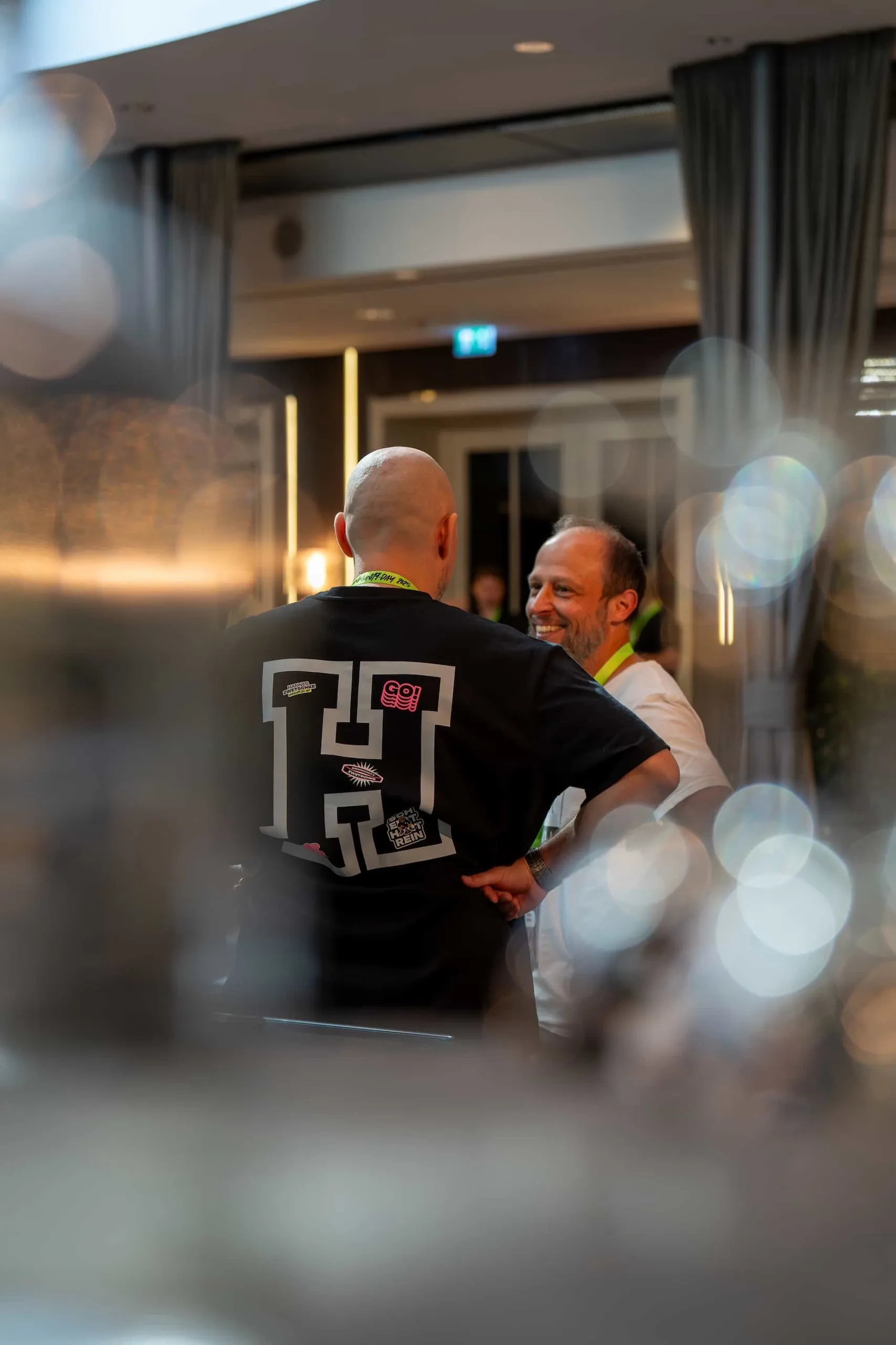 Hannes Zwetschke begrüßt einen Gast beim Hannes Zwetschke Community-Day 2025. Auf dem Rücken des schwarzen Shirts ein großes weißes „H", übersät mit Event-Stickern.