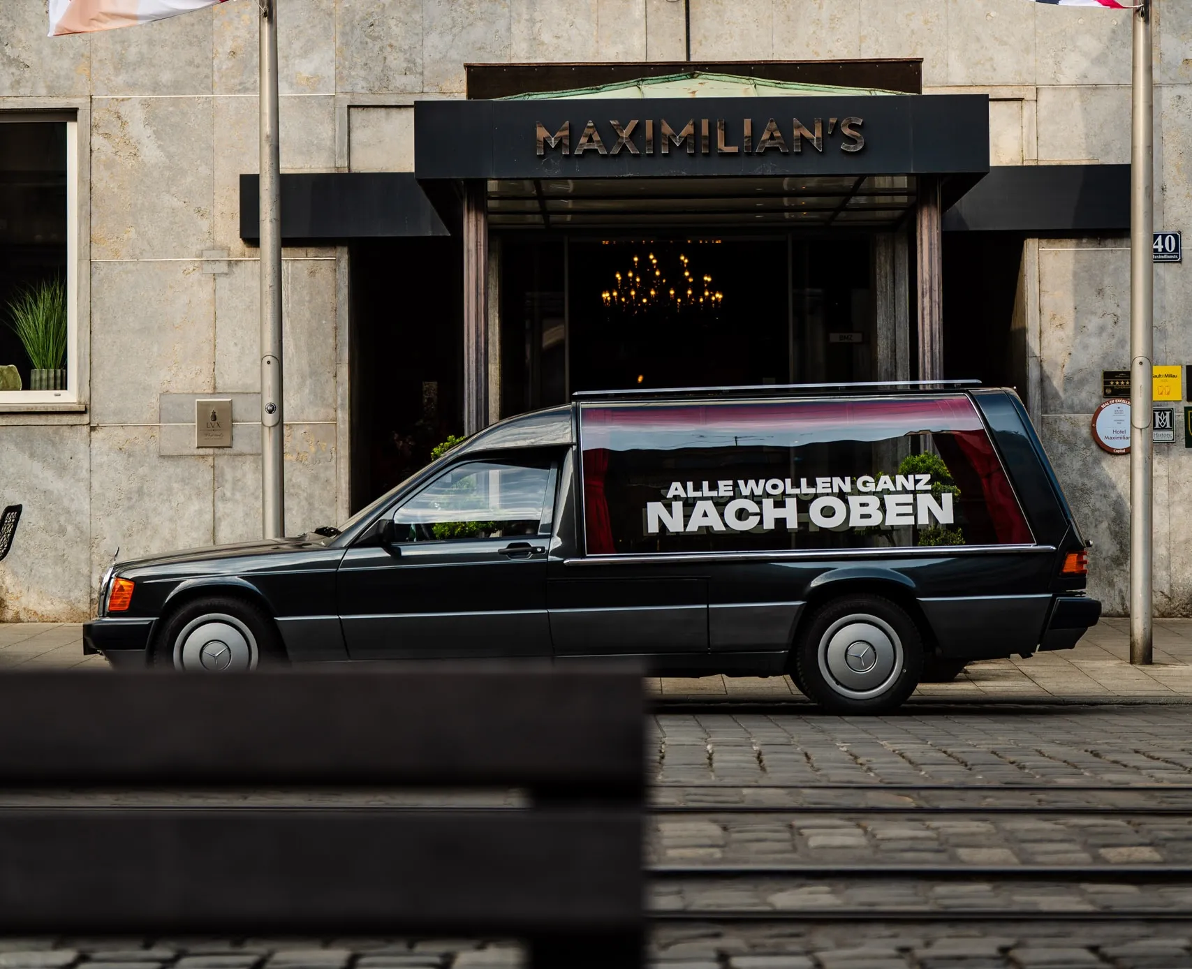 Hannes Zwetschkes eigener Leichenwagen, ein schwarzer Mercedes mit der Aufschrift „Alle wollen ganz nach oben", parkt vor dem Hotel Maximilian's in Augsburg, dem Veranstaltungsort des Zwetschke Community-Day 2025.