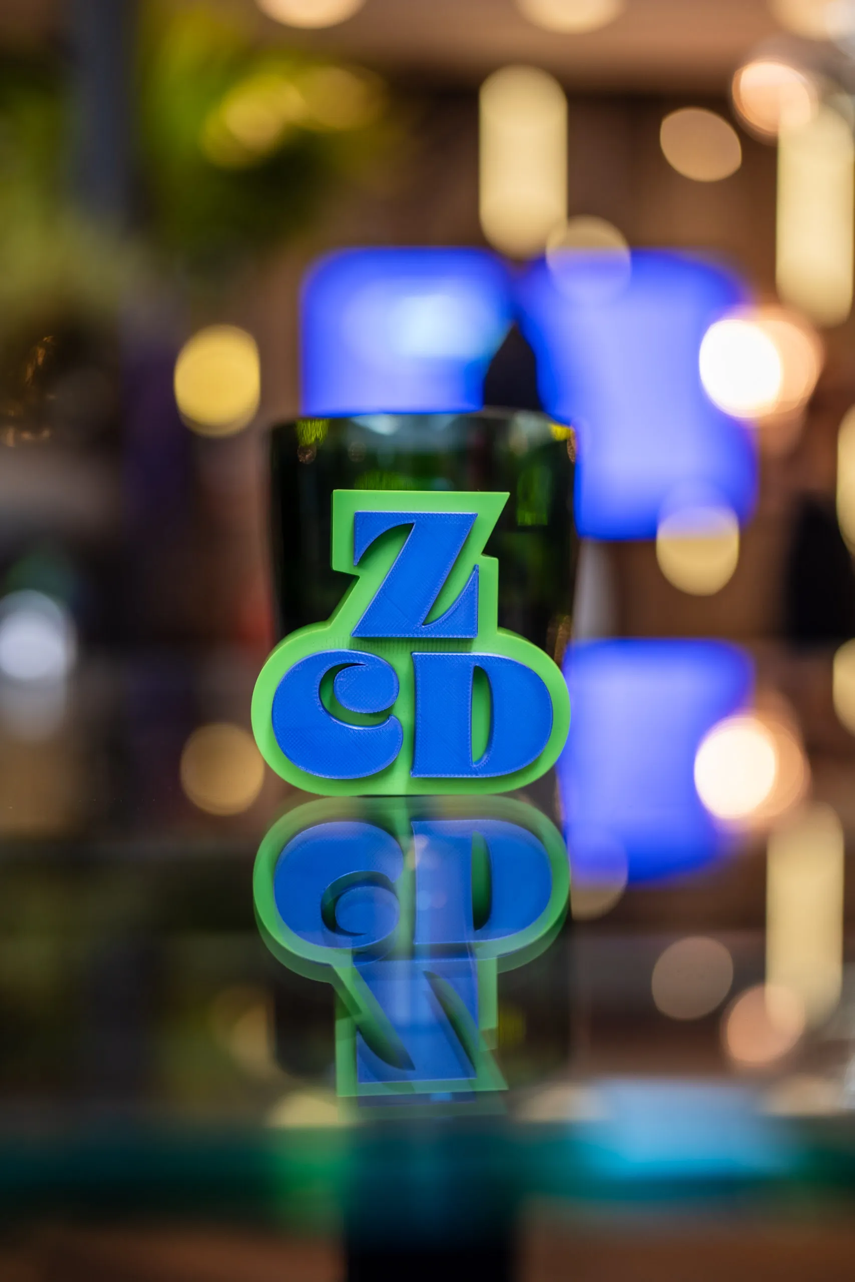 3D-gedrucktes ZCD-Logo in Blau und Neongrün auf einer spiegelnden Glasfläche, dahinter unscharfe Lichter des Zwetschke Community Day 2026.