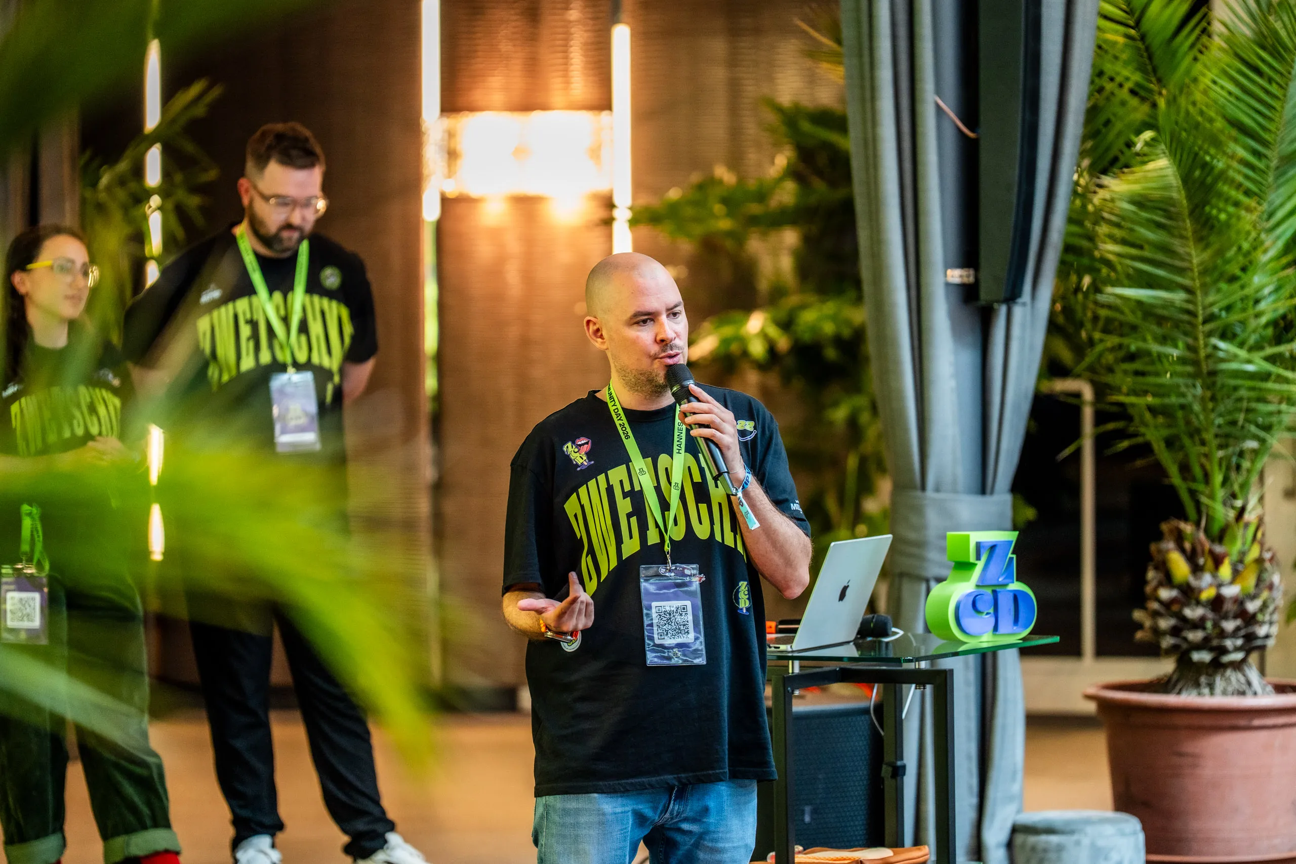 Hannes Zwetschke moderiert mit Mikrofon seinen Community Day 2026 im Hotel Maximilian's in Augsburg, dahinter zwei Teammitglieder in ZWETSCHKE-Shirts.
