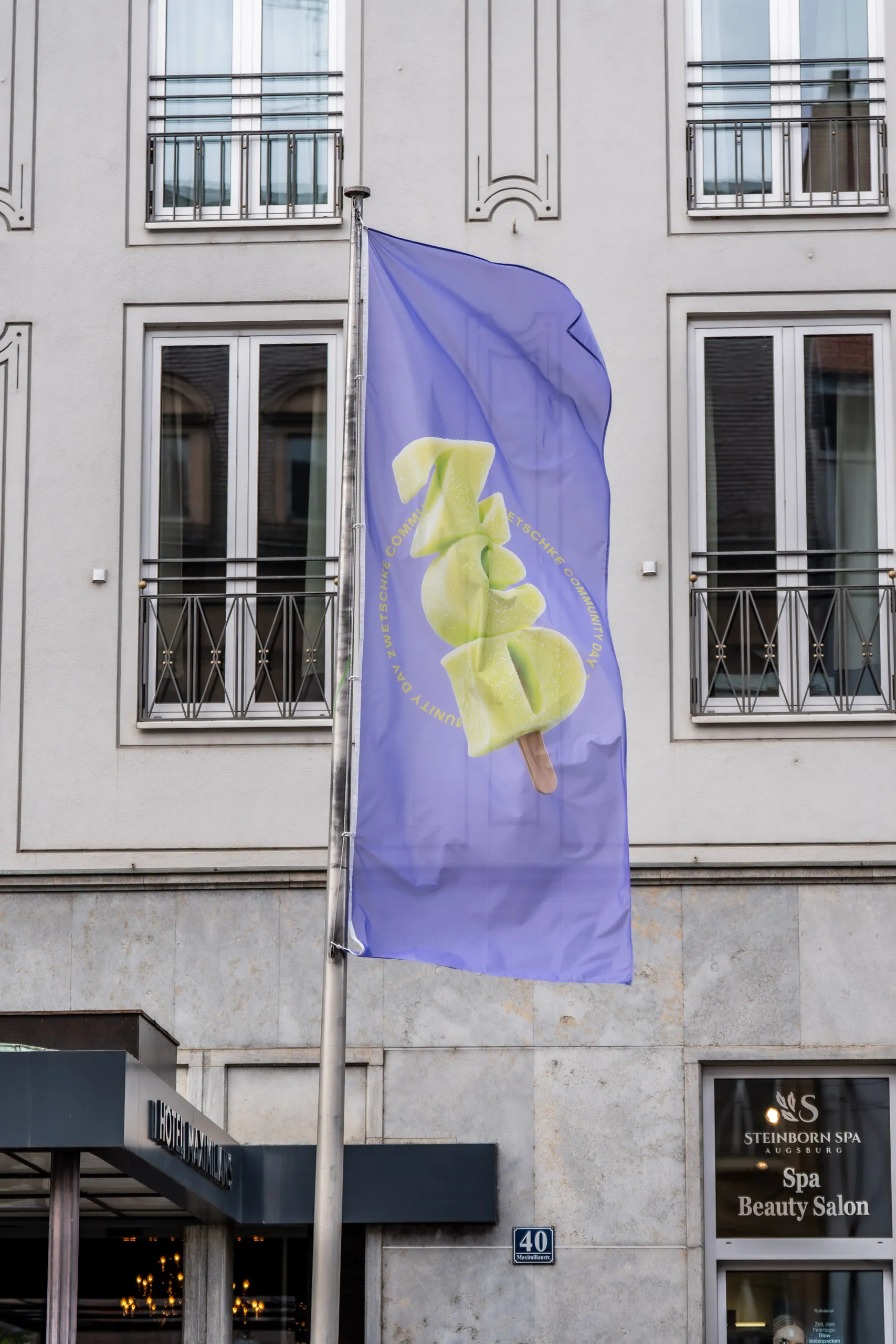 Violette Eventfahne mit dem Logo des Zwetschke Community Day 2026 als grünes Eis am Stiel, vor dem Hotel Maximilian's in der Maximilianstraße in Augsburg.