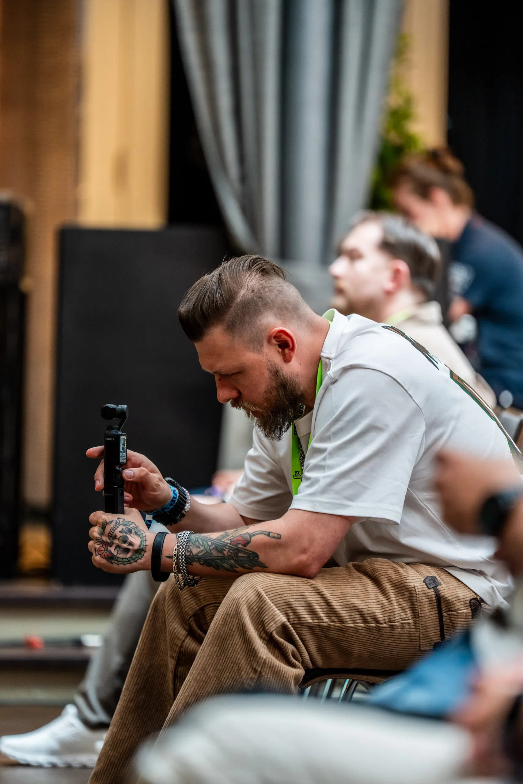 Gast mit Tattoos und Vollbart sitzt im Publikum des Zwetschke Community Day 2026 und filmt mit einer Pocket-Kamera.