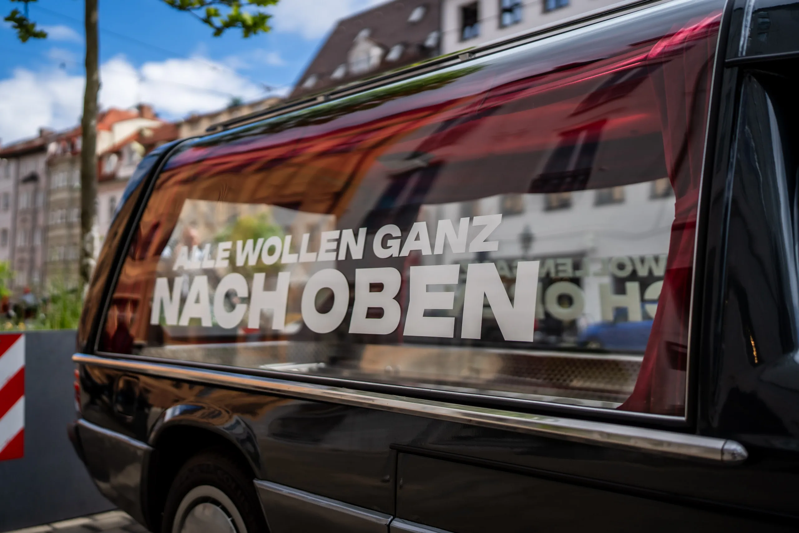 Detailaufnahme der Heckscheibe von Hannes Zwetschkes Leichenwagen mit dem Schriftzug „Alle wollen ganz nach oben", darin spiegelt sich die Maximilianstraße.