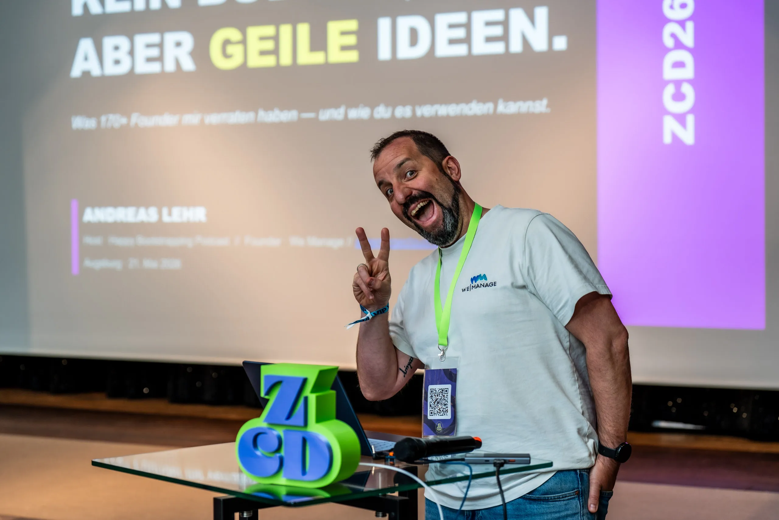 Andreas Lehr zeigt lachend ein Peace-Zeichen vor seiner Titelfolie „Kein Budget, aber geile Ideen" beim Zwetschke Community Day 2026 in Augsburg.