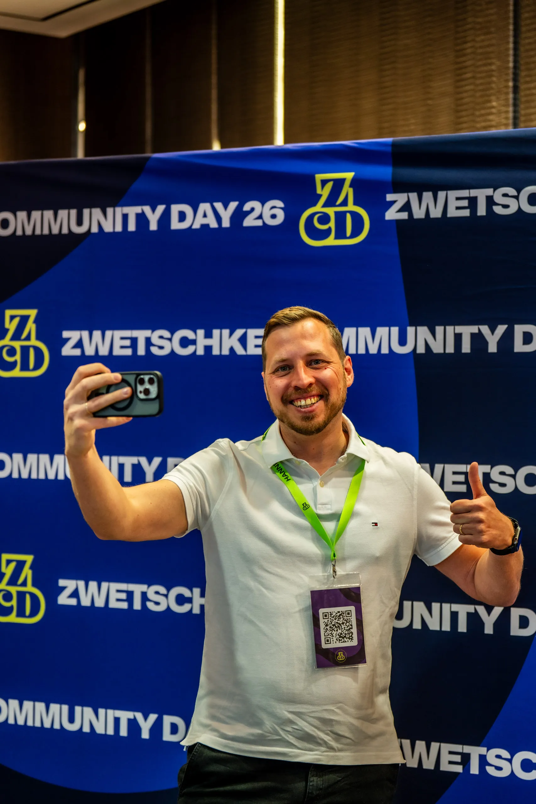 Gast macht ein Selfie mit erhobenem Daumen vor der blauen ZCD26-Fotowand beim Zwetschke Community Day 2026 in Augsburg.