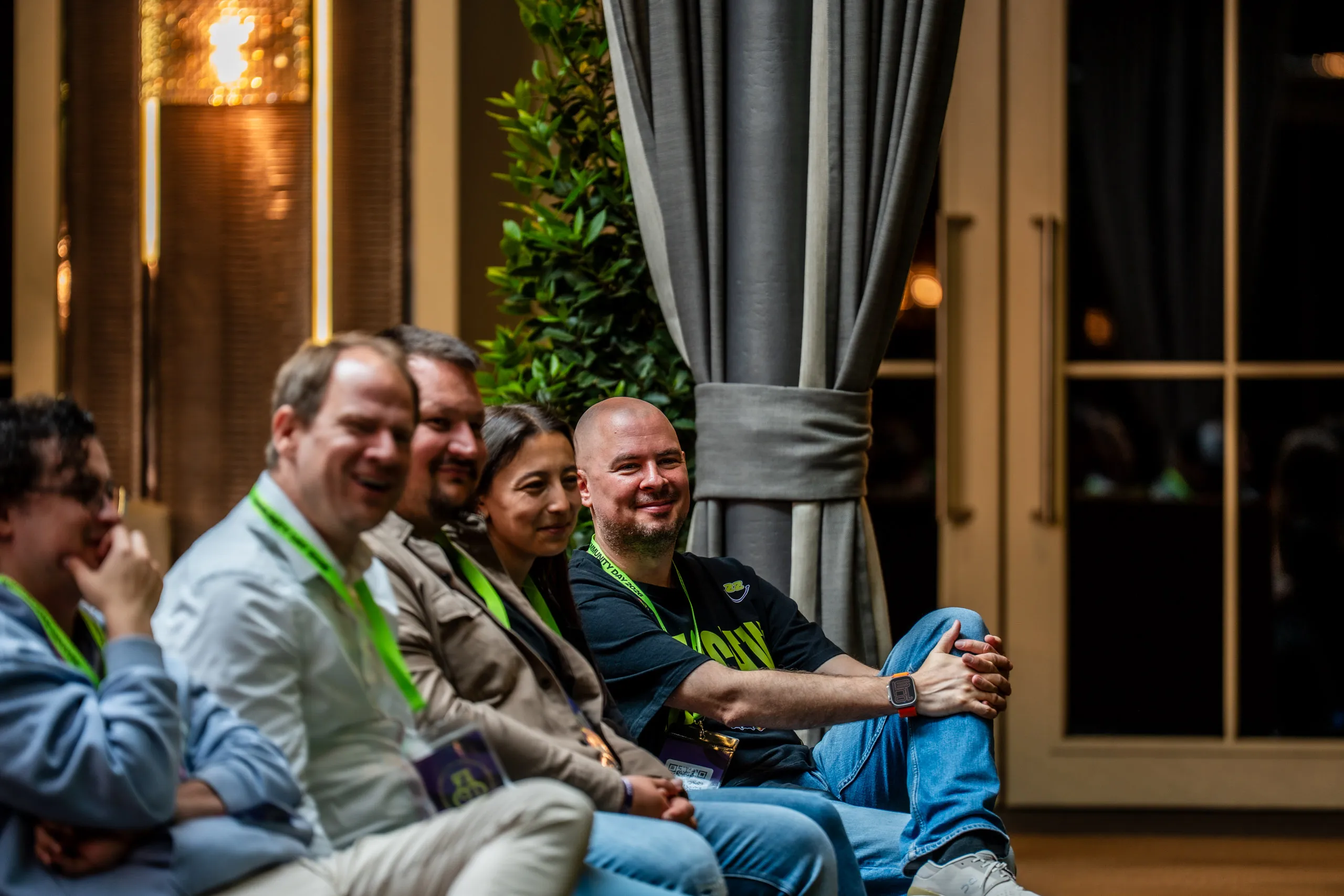 Hannes Zwetschke sitzt entspannt in einer Reihe lachender Gäste beim Zwetschke Community Day 2026 im Hotel Maximilian's in Augsburg, im warmen Licht.