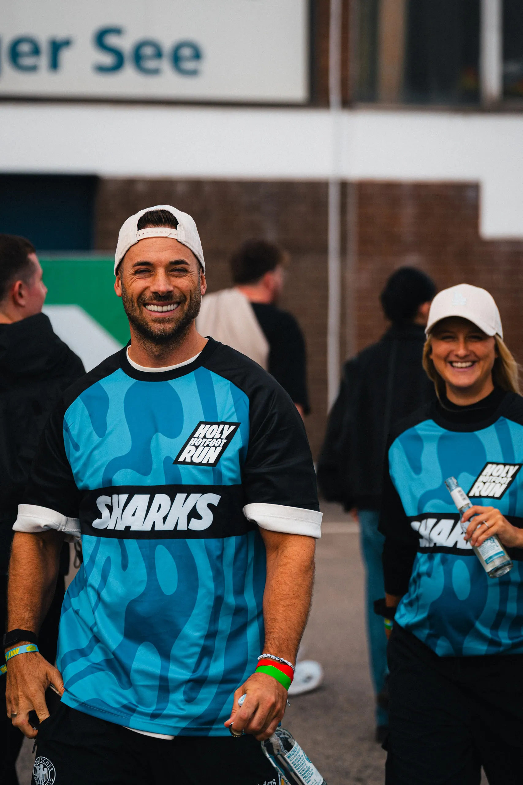 A laughing man in a white backwards cap wears a blue "Sharks" jersey with the HOLY Hotfoot Run logo; behind him a woman in the same team jersey.