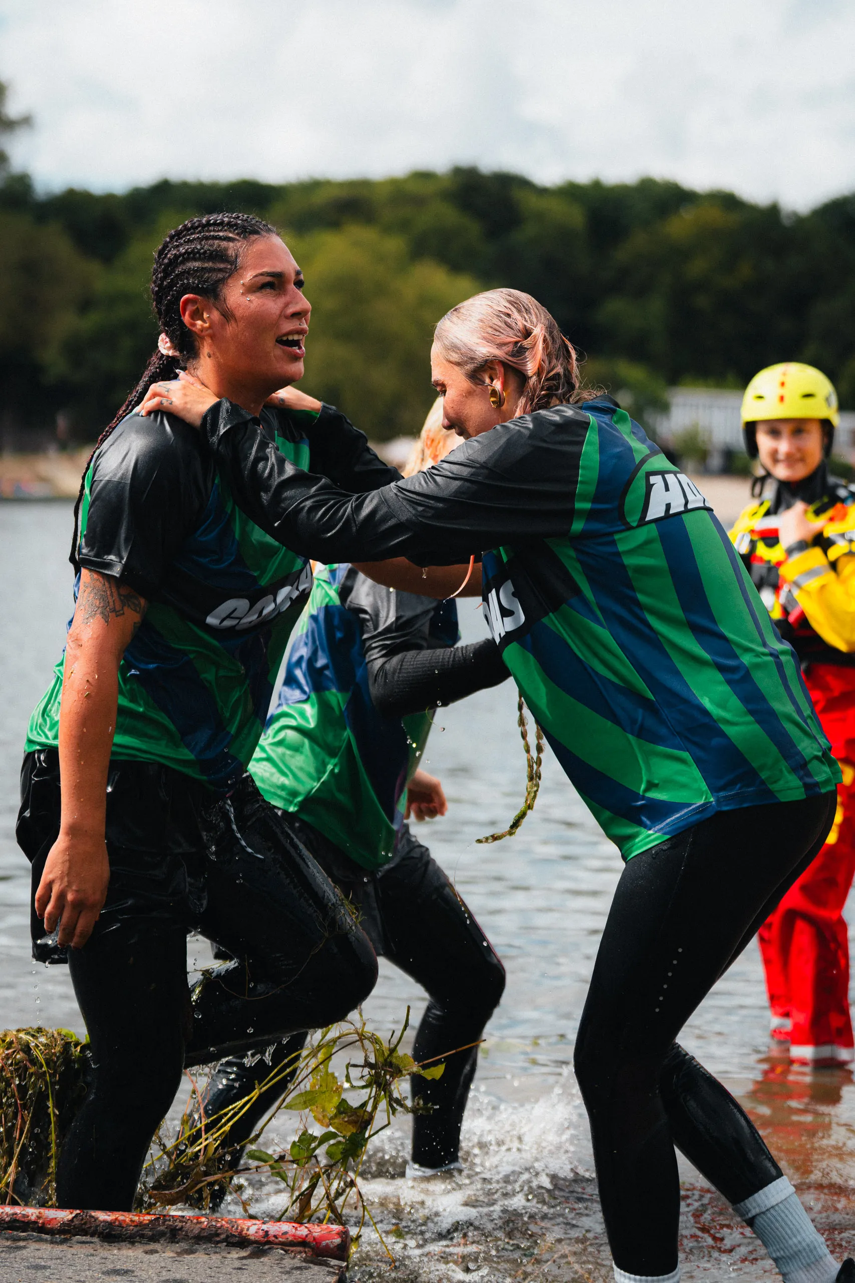 Two soaked runners in Cobras jerseys climb out of the water, one steadying the other, with a water-rescue crew member in a yellow helmet at the right.