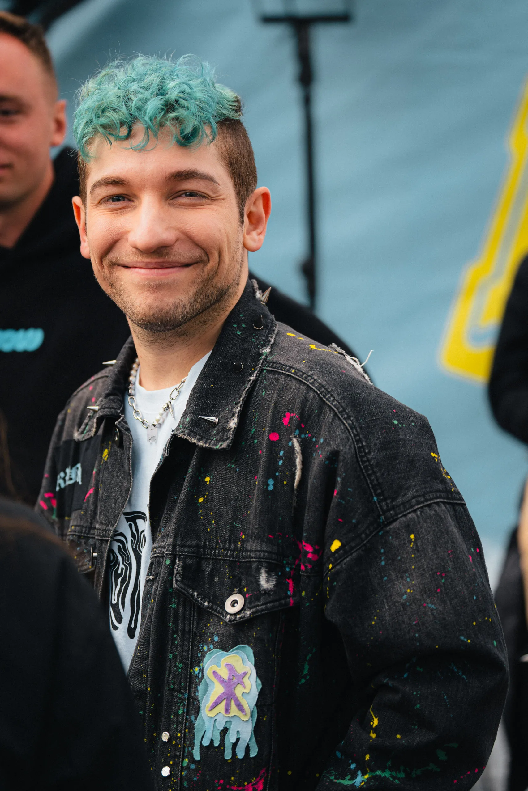 Rezo in a black denim jacket covered in colourful paint splatters, studs and patches, a pale blue event banner behind him.