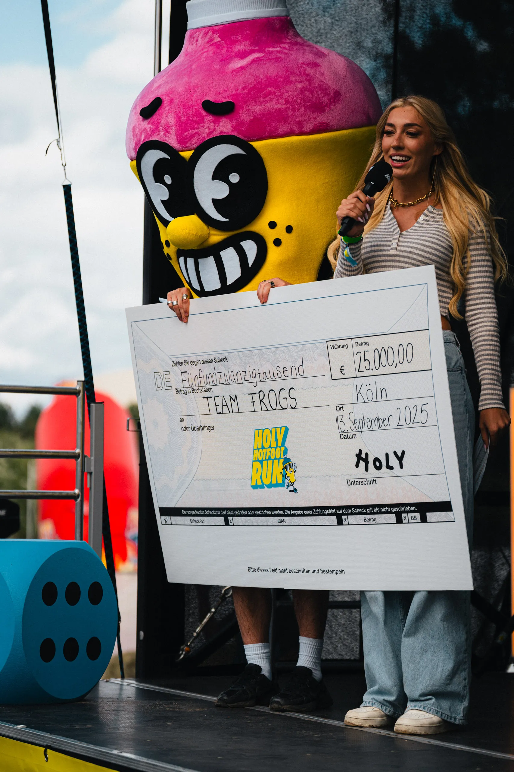 A host with a microphone and a mascot in a yellow cartoon-figure costume hold up an oversized cheque for €25,000 made out to "Team Frogs", dated Cologne, 13 September 2025.