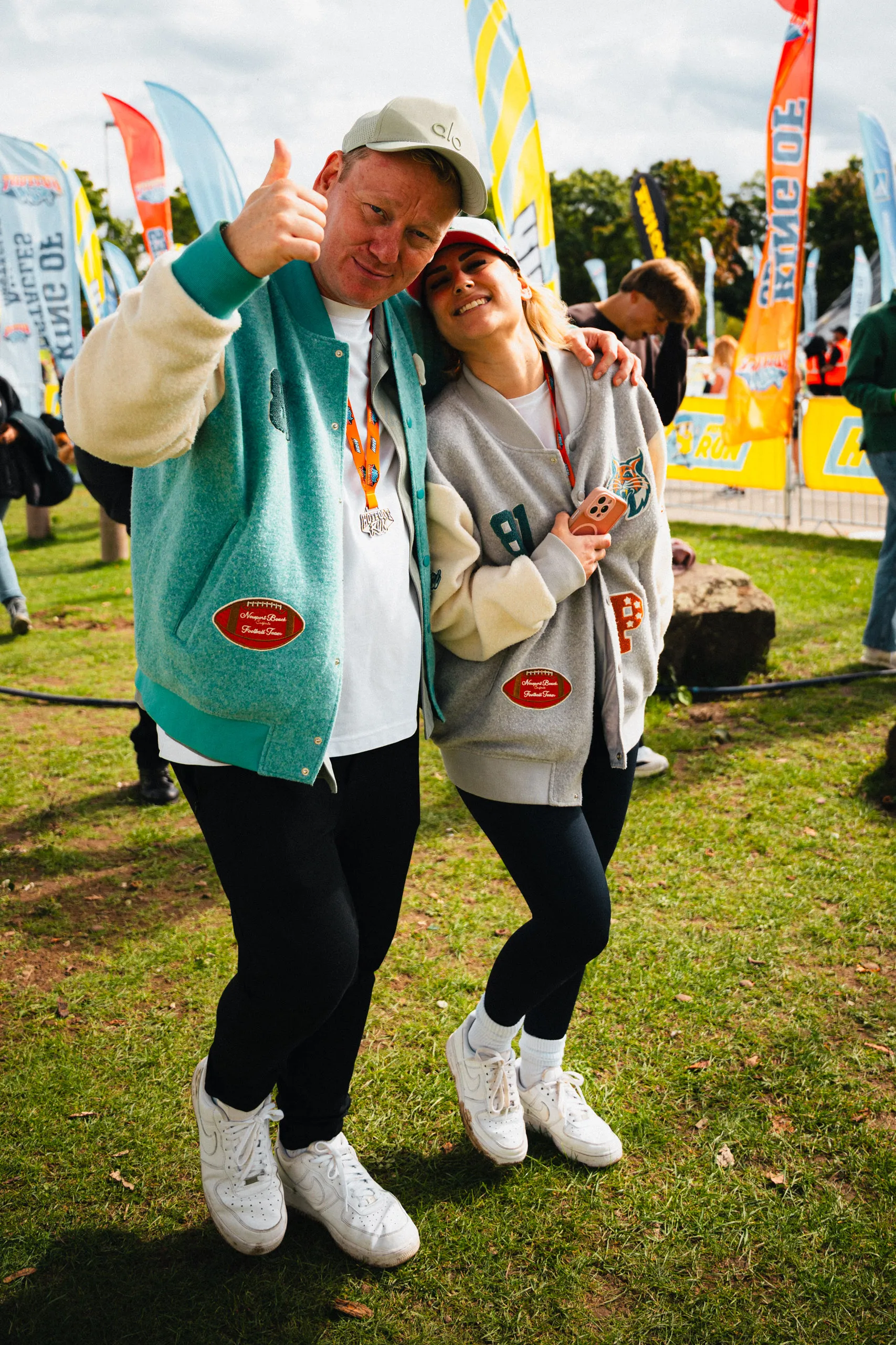 Knossi in a mint PARI varsity jacket gives a thumbs up, a woman in a grey PARI varsity jacket leaning against him; both wear orange event lanyards, with Hotfoot Run beach flags behind them.