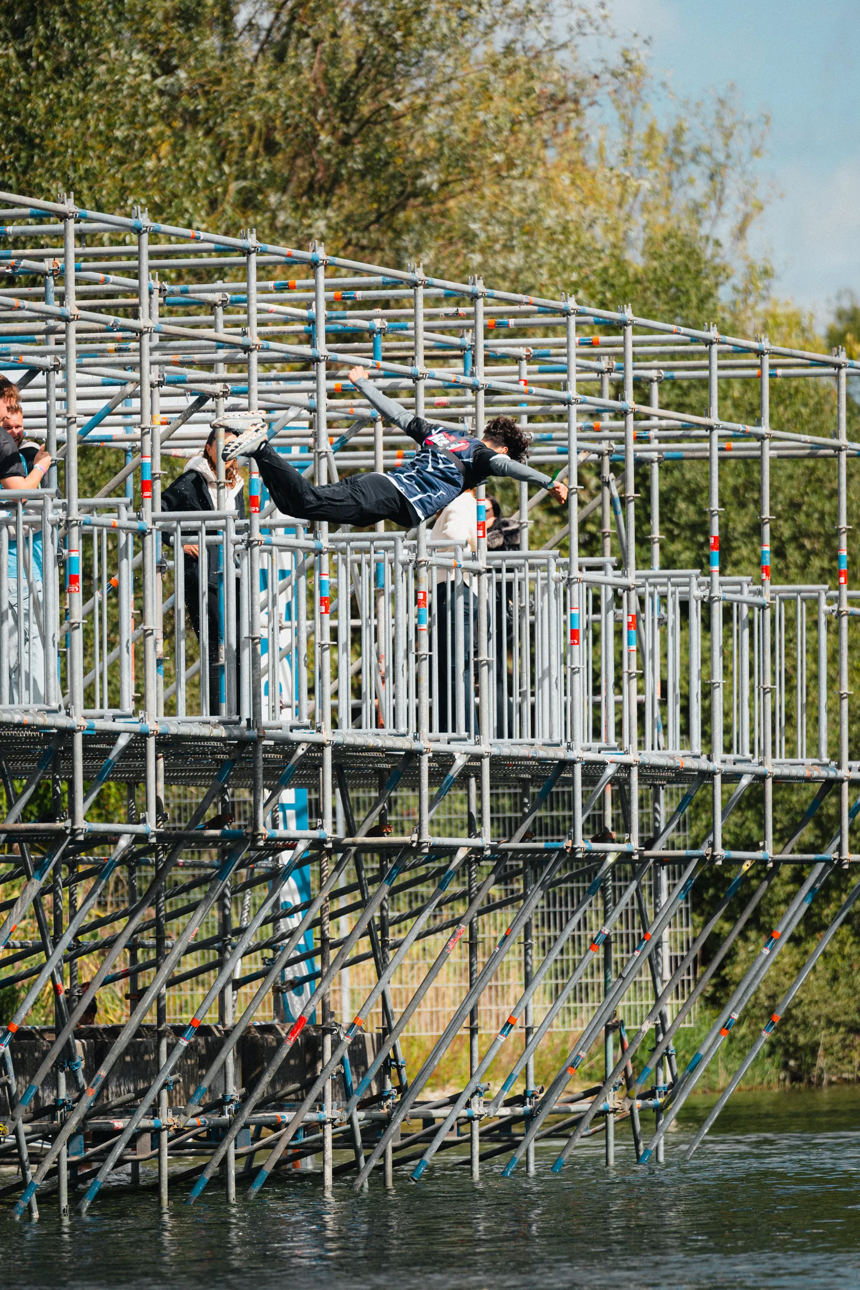 A person jumps with arms outstretched from a scaffolding obstacle set up directly over the water; other participants are waiting on the platform behind it.