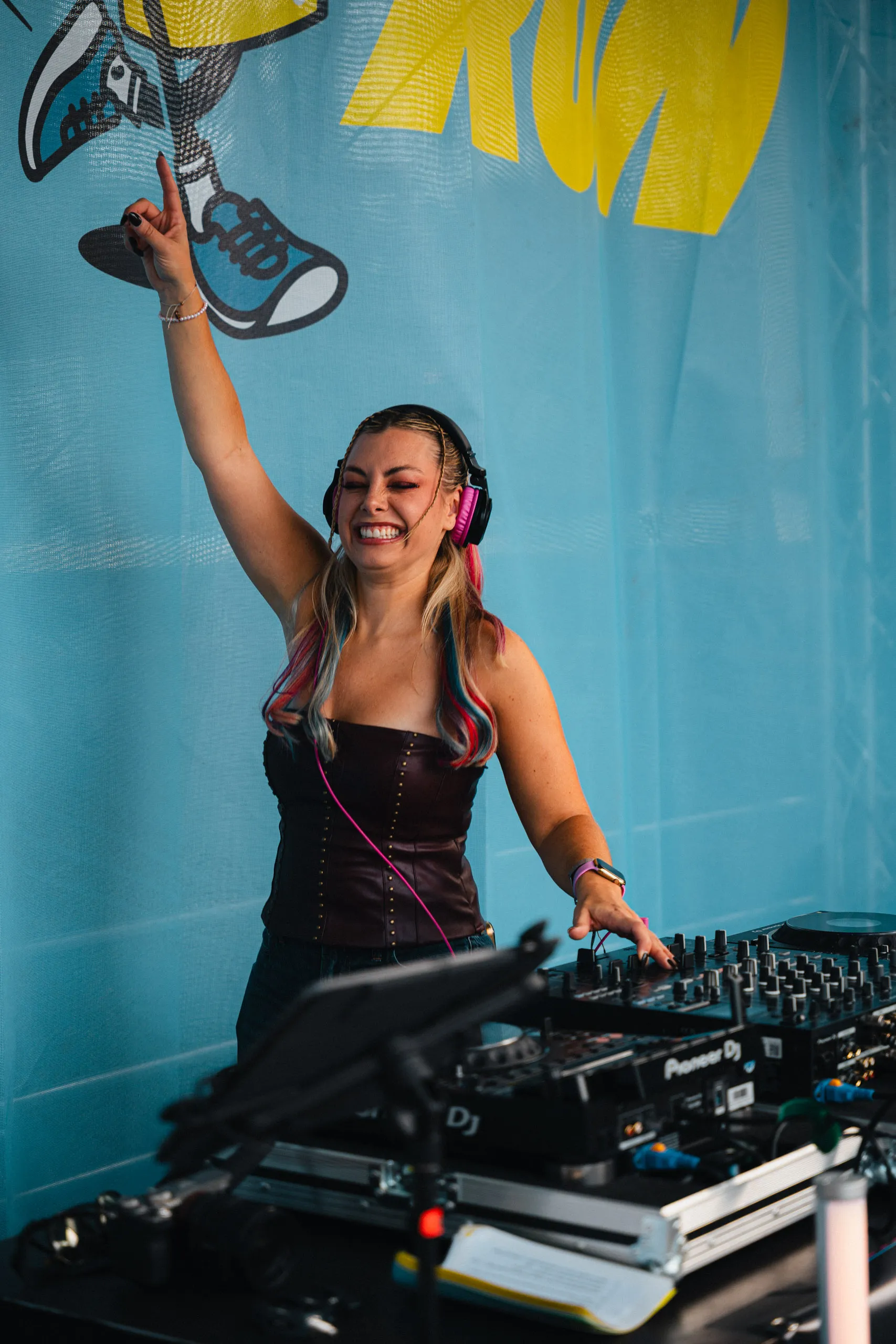 Chany Dakota DJs at a Pioneer setup, headphones on, one arm raised as she laughs, with a pale blue Hotfoot Run banner behind her.