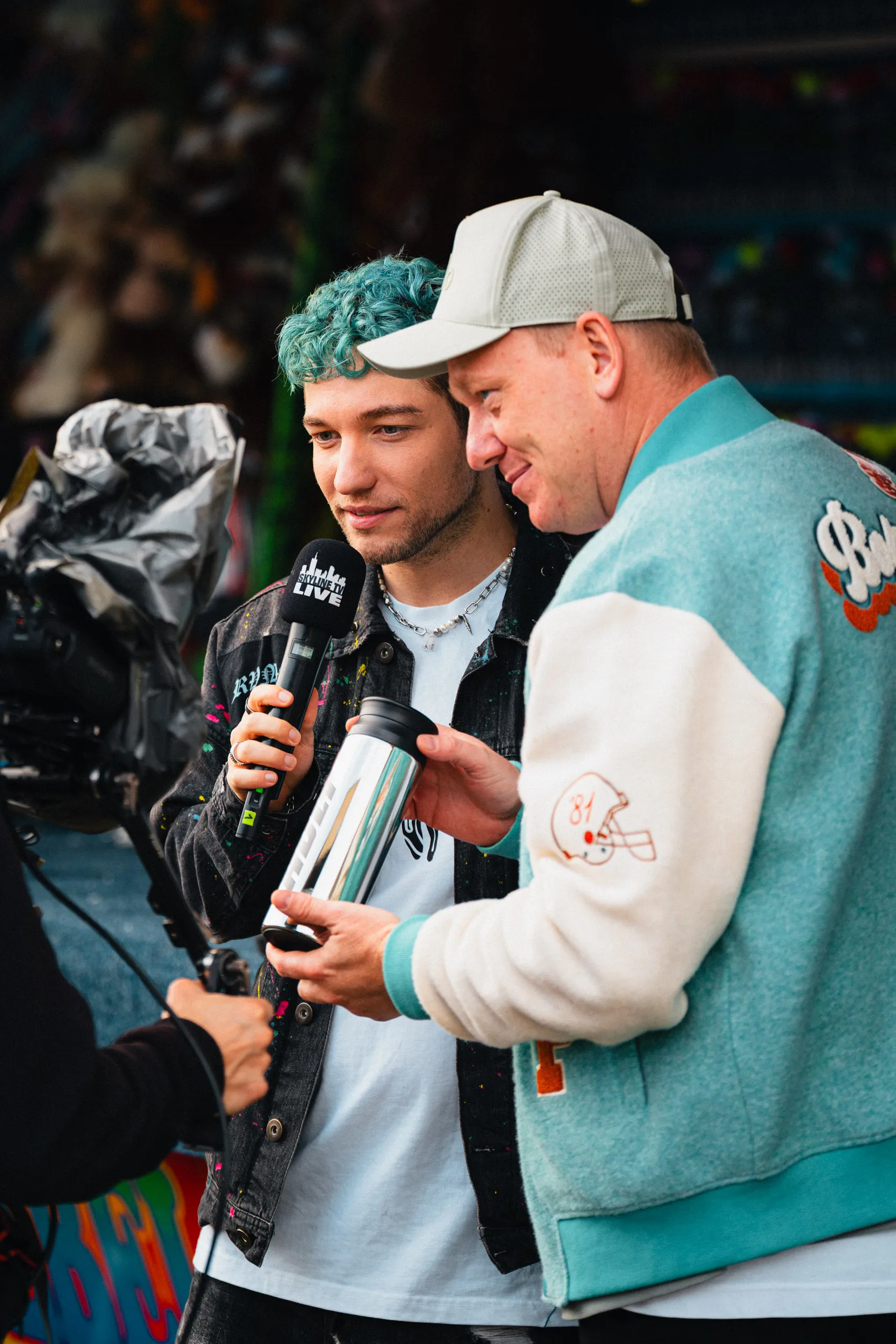 Rezo mit türkisfarbenen Locken hält ein Mikrofon mit der Aufschrift „Skyline TV Live", daneben Knossi in mintgrüner Collegejacke mit einer silbernen Trinkflasche in der Hand. Links im Bild eine Kamera mit Regenschutz.