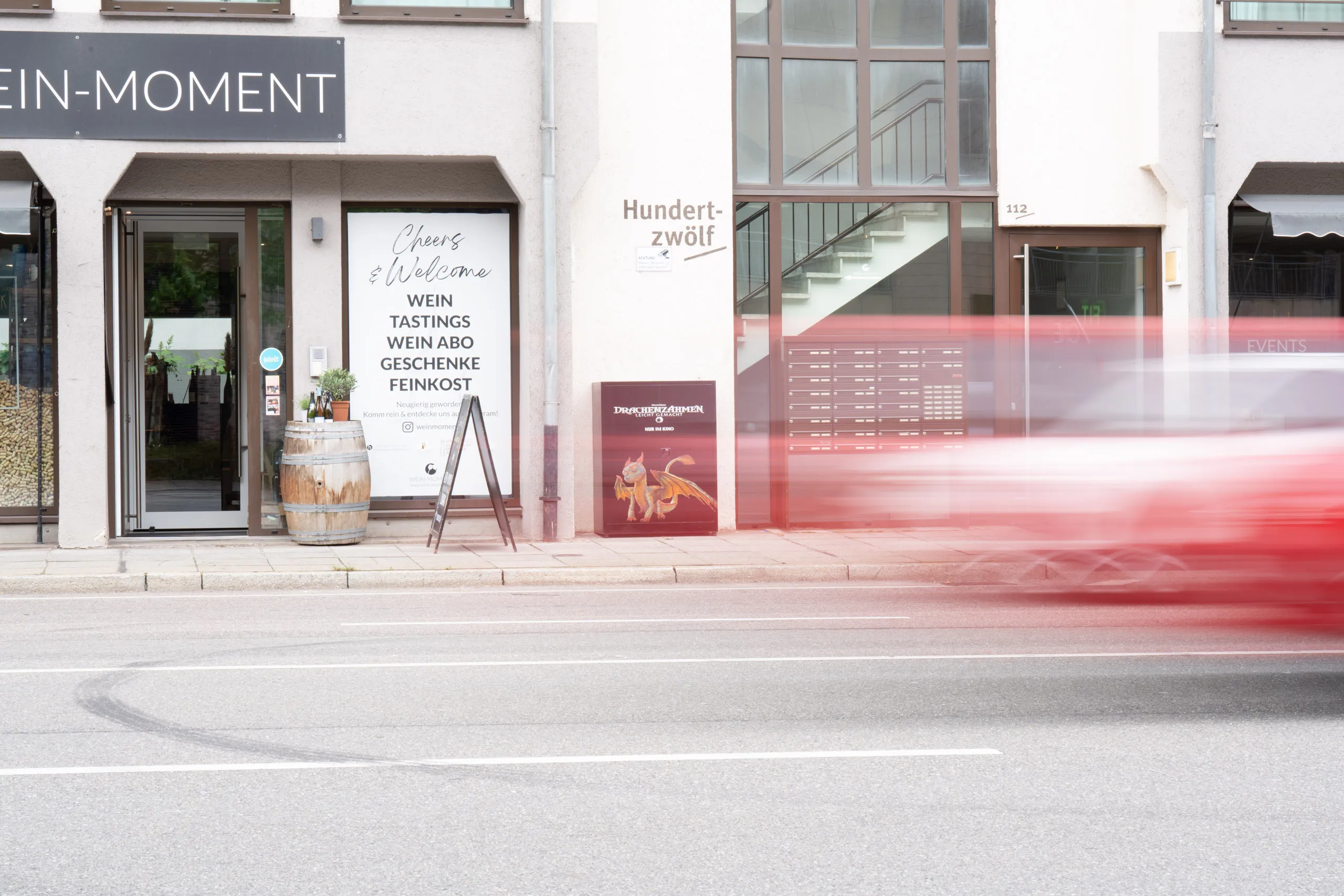 Street view in Stuttgart: a black electrical cabinet with dragon artwork stands against the wall beside a wine shop, a red vehicle blurring past on the right.