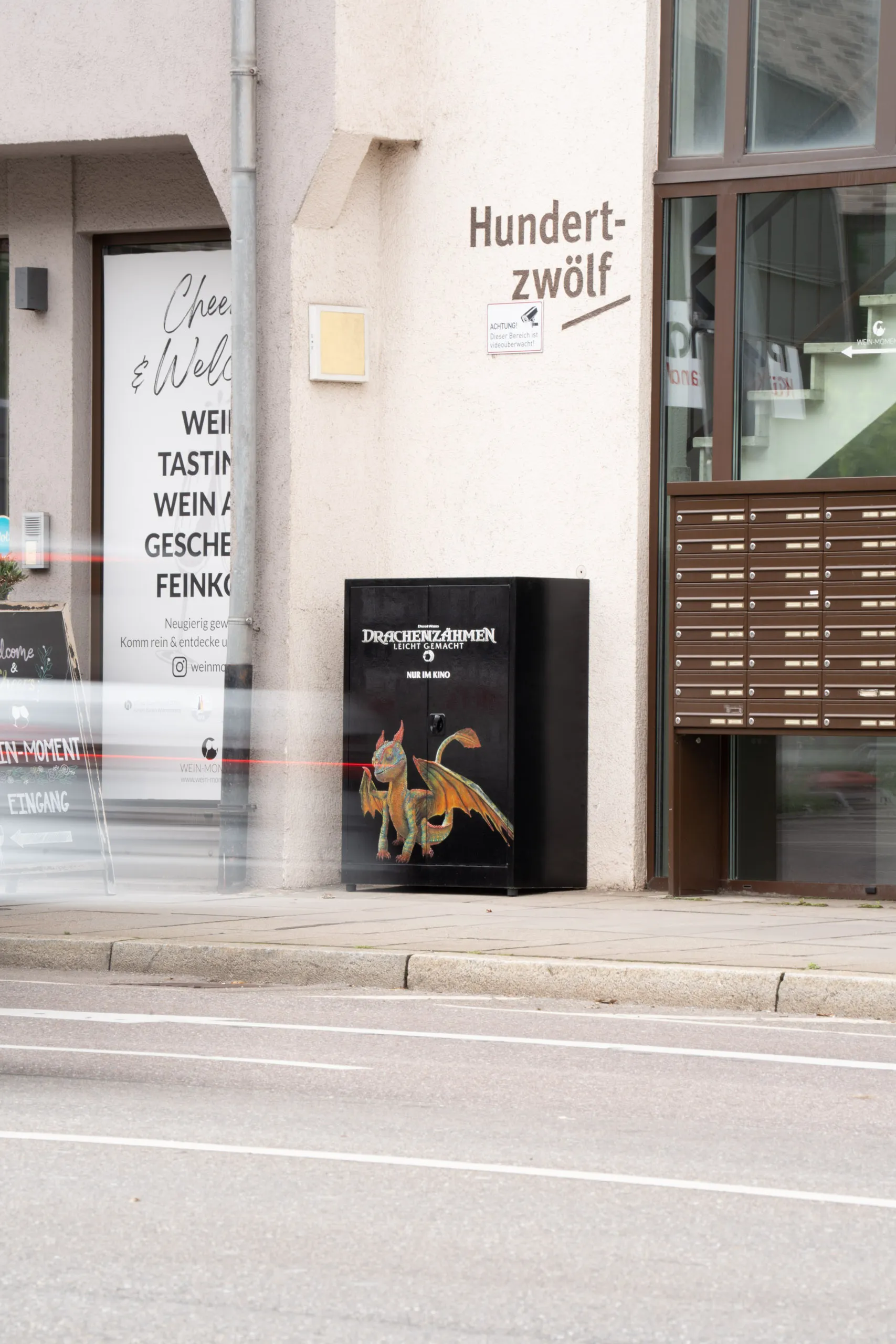 The dragon-painted electrical cabinet stands against a pale wall next to a bank of letterboxes, with red and white light trails from a passing vehicle streaking across the left of the frame.