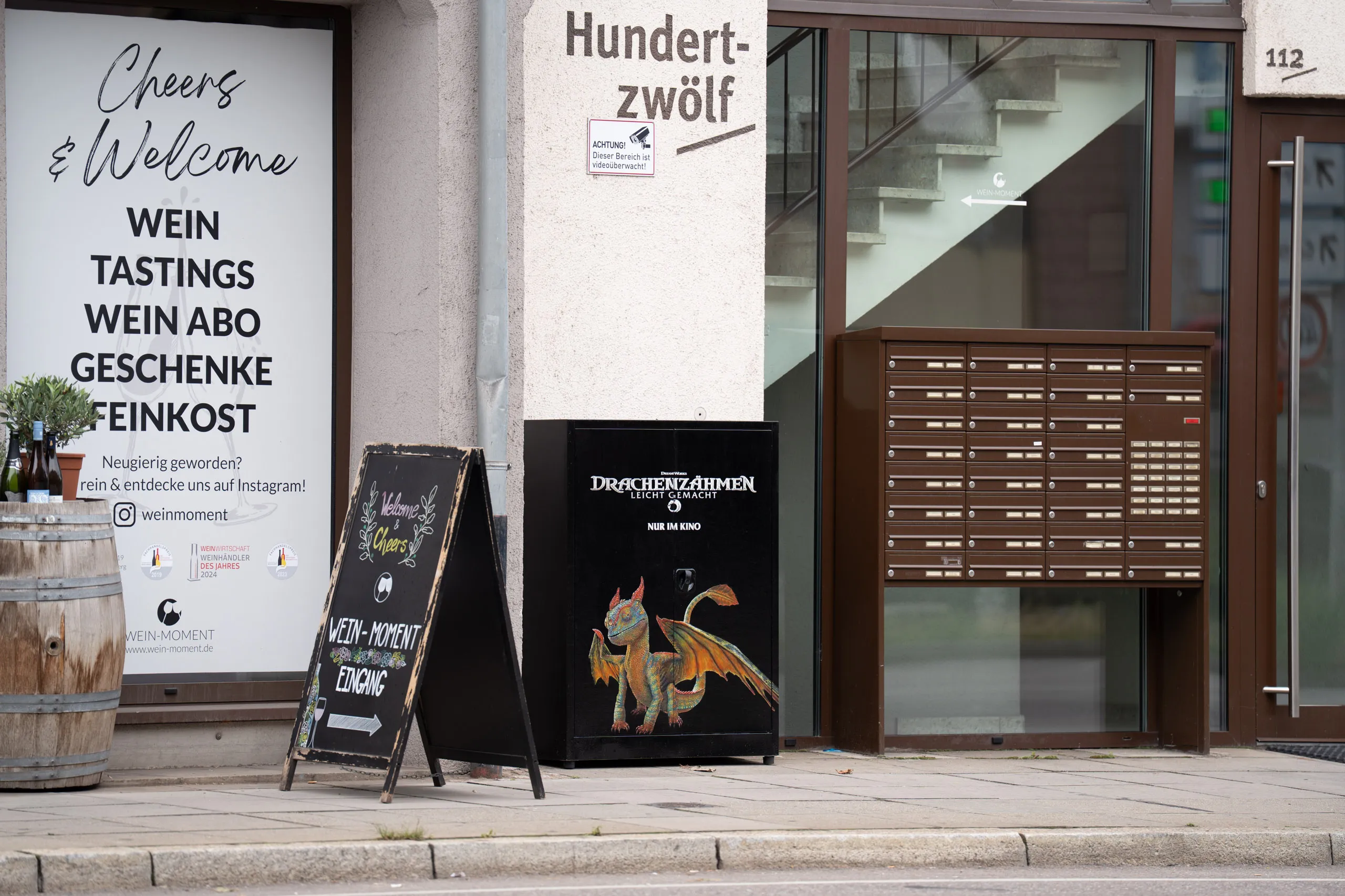 The dragon-painted electrical cabinet sits between a pavement chalkboard and a bank of letterboxes, a wine barrel with an olive plant and bottles in front of the shop window to the left, and the building name "Hundertzwölf" above.
