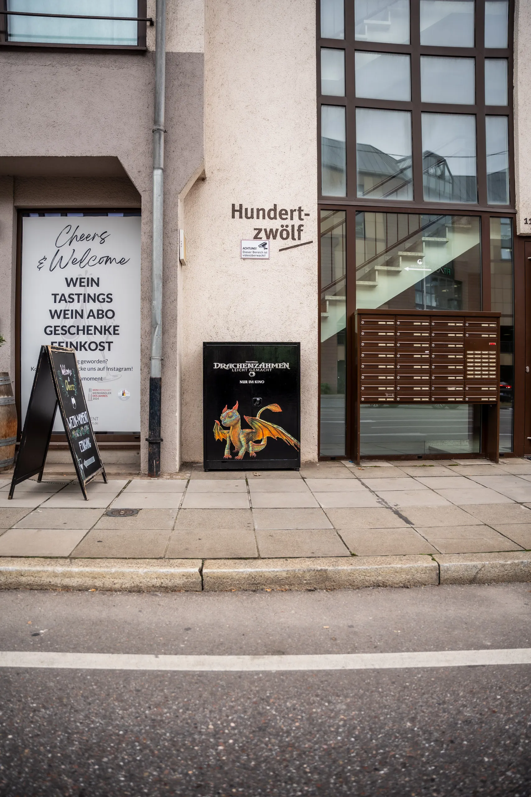 A wide shot of the building facade: the dragon-painted electrical cabinet sits small between a downpipe and the letterboxes, an empty stretch of pavement in front.