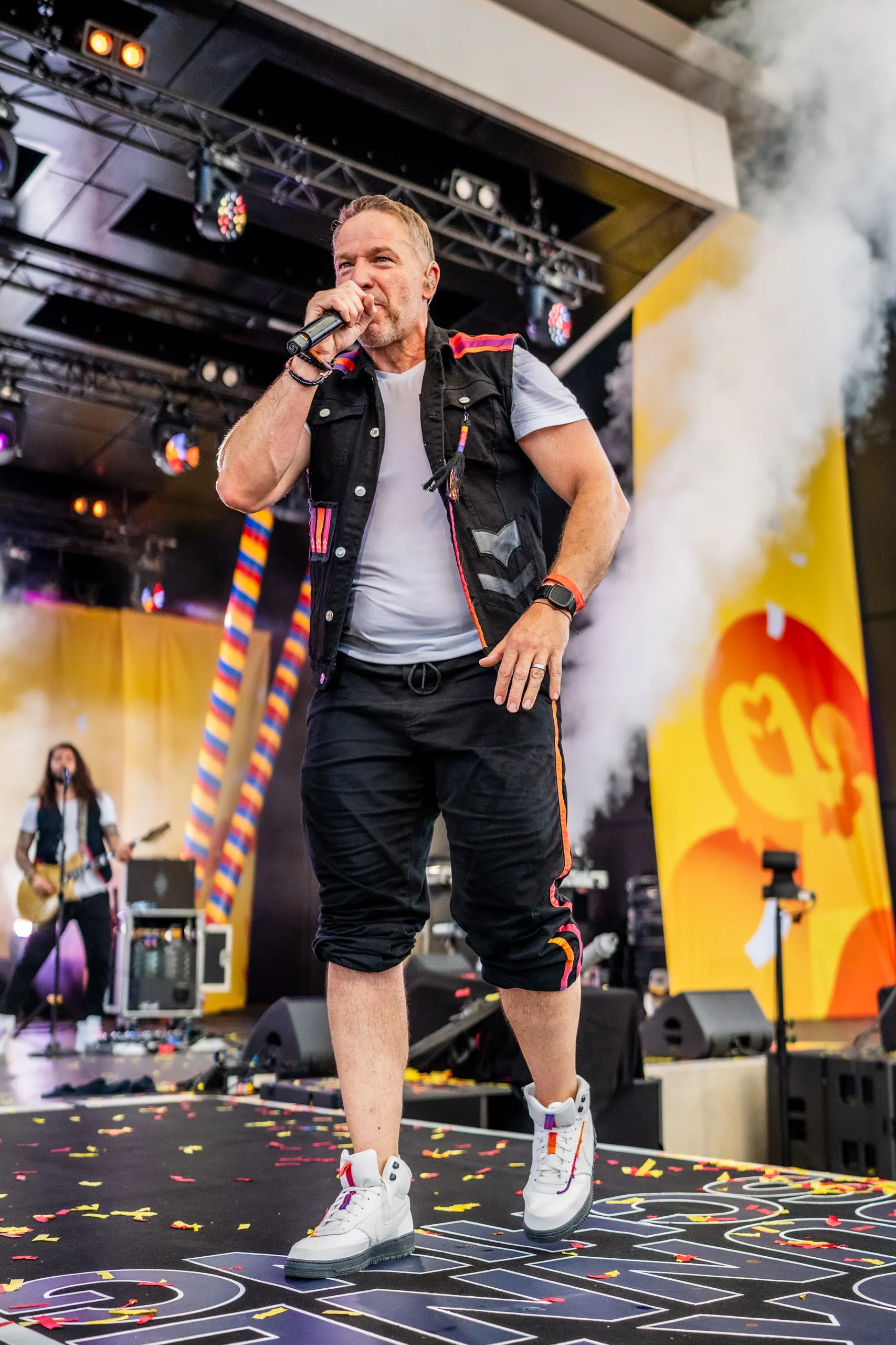 Patrick Lück, frontman of Höhner, with a microphone on the stage catwalk at Jeck im Sunnesching, a guitarist and drifting smoke behind him.