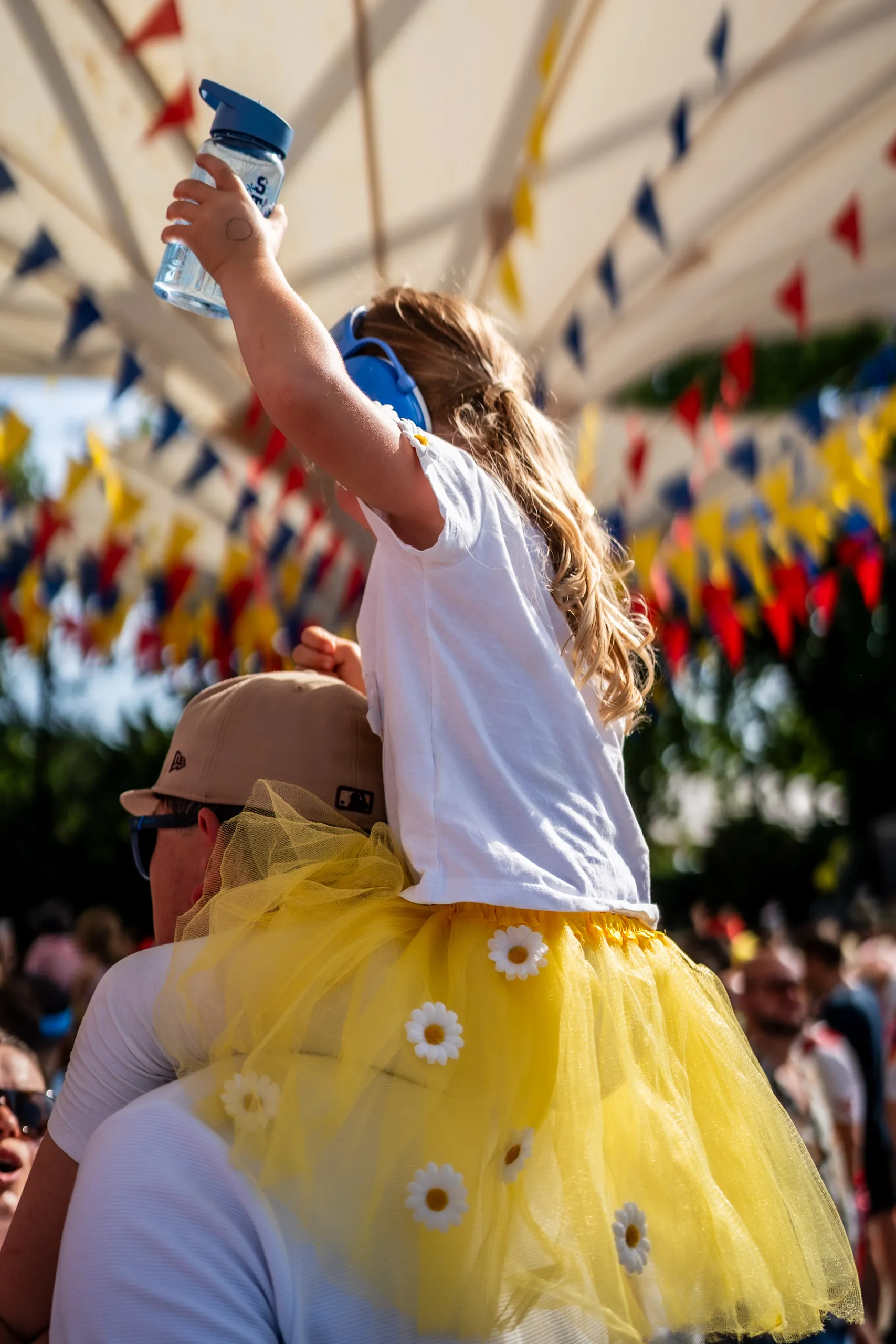 A girl in blue ear defenders and a yellow tutu sits on her father’s shoulders, holding a water bottle up in the air, with colourful bunting behind her.