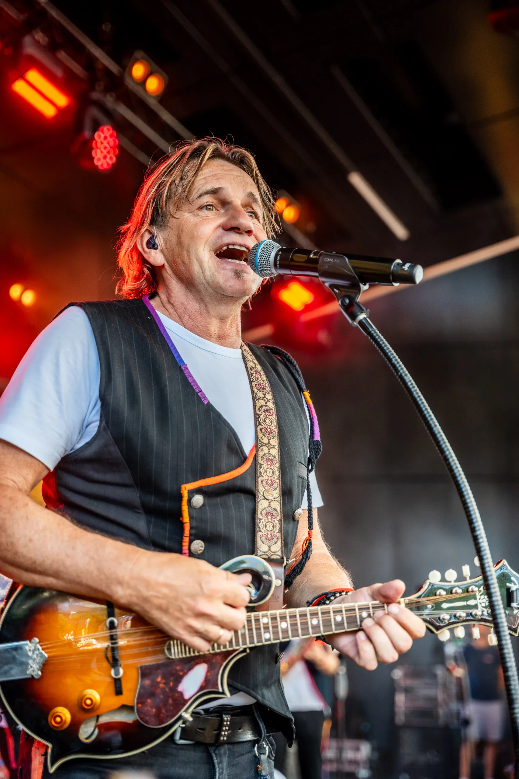 Jens Streifling of Höhner singing into the microphone while playing a mandolin, lit in red stage light.