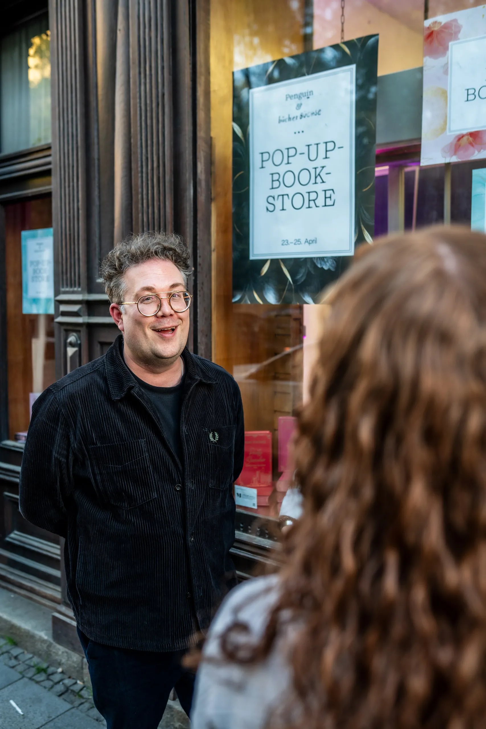 Thomas Sachsenmaier outside the pop-up book store window talking with a visitor, the poster with the 23–25 April dates behind the glass.