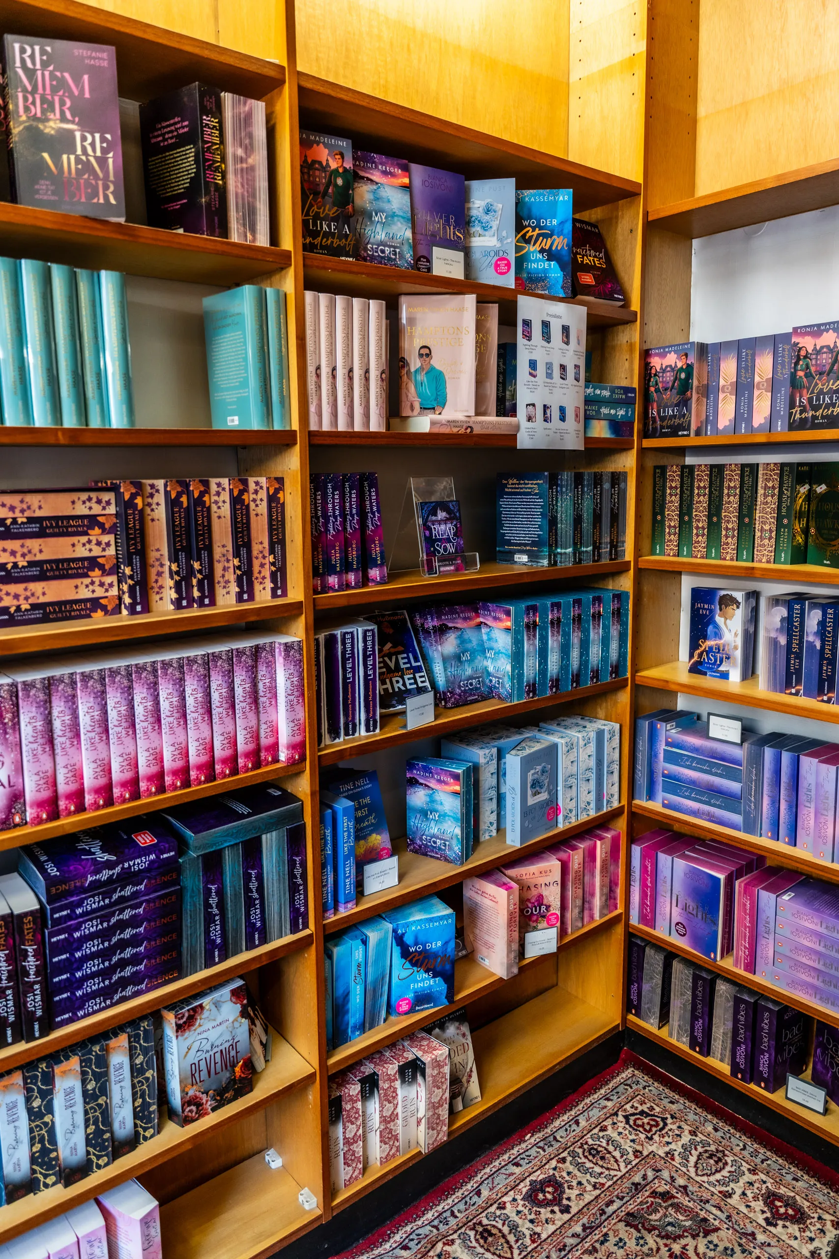 Densely packed wooden shelves of romance and romantasy titles at the pop-up book store, a Persian rug in front.