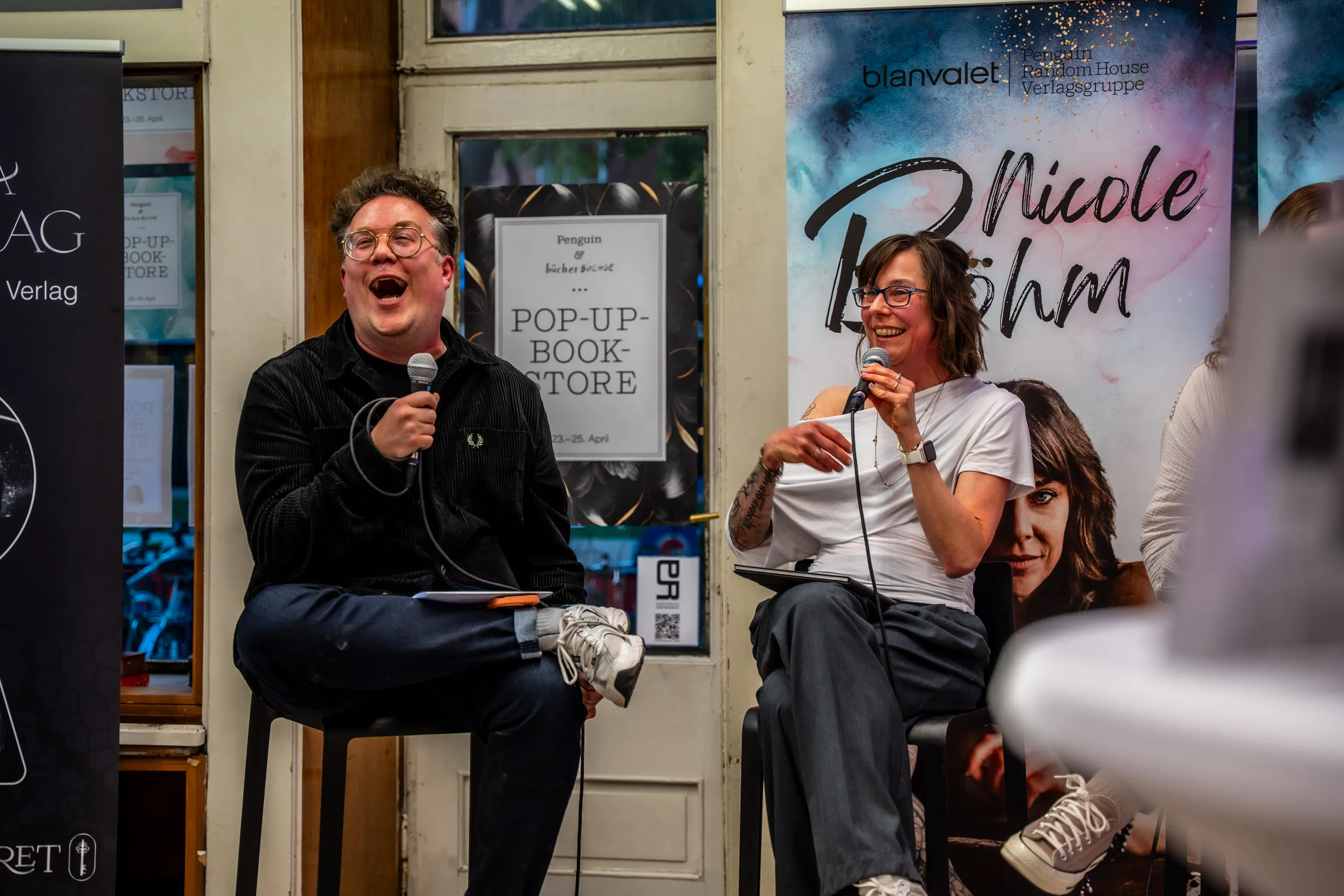 Thomas Sachsenmaier laughing with a microphone on his bar stool, Nicole Böhm beside him in front of her author banner during the talk at the pop-up book store.