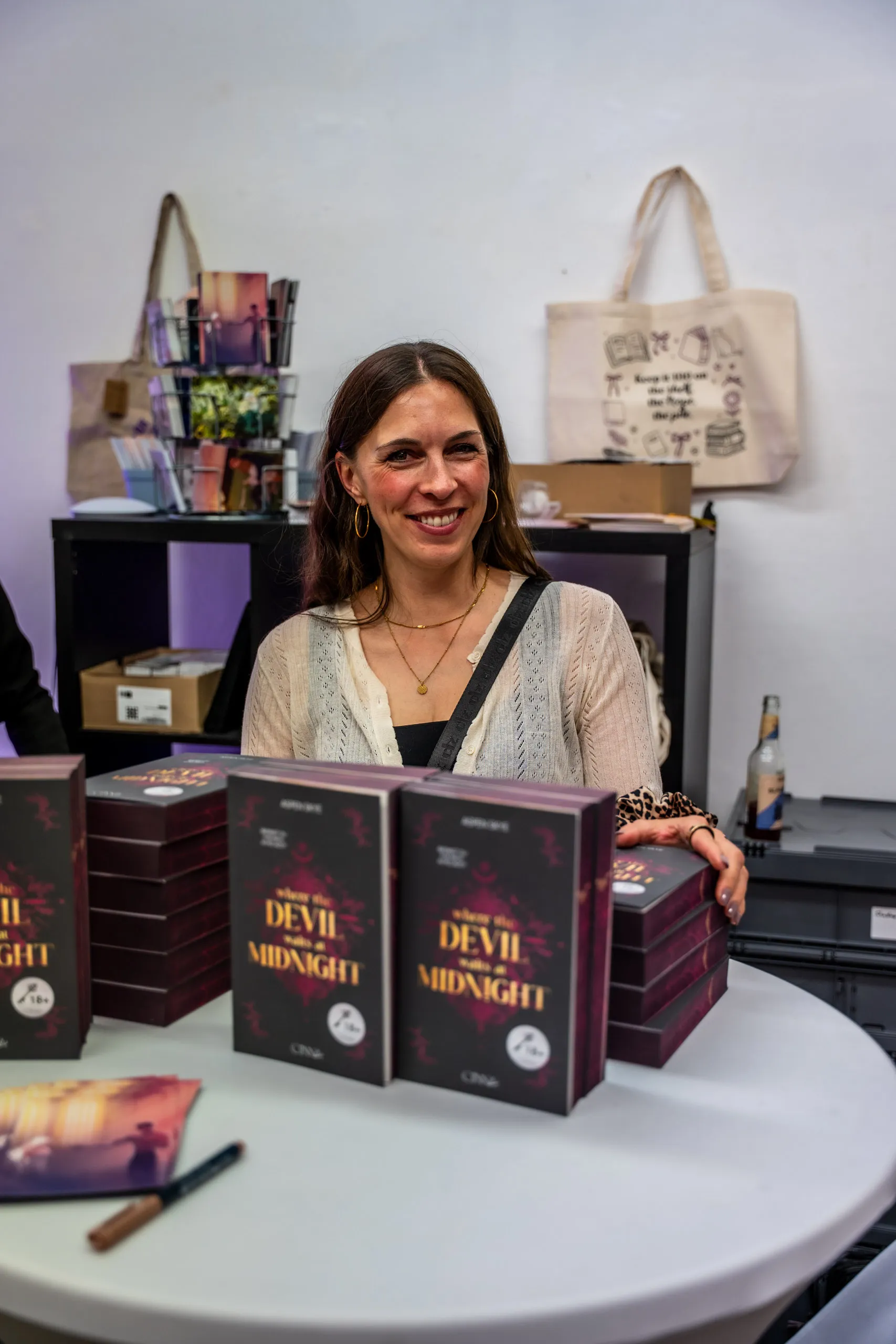 Aspen Skye at the signing table behind stacks of her novel "Where the Devil Waits at Midnight", pop-up book store merch bags behind her.