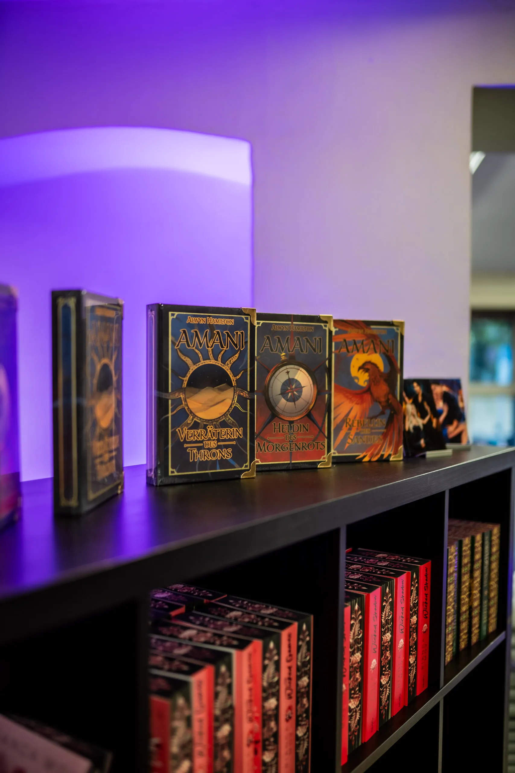 Collector’s editions of Alwyn Hamilton’s Amani trilogy on a shelf in purple light, rows of further sprayed-edge editions below.