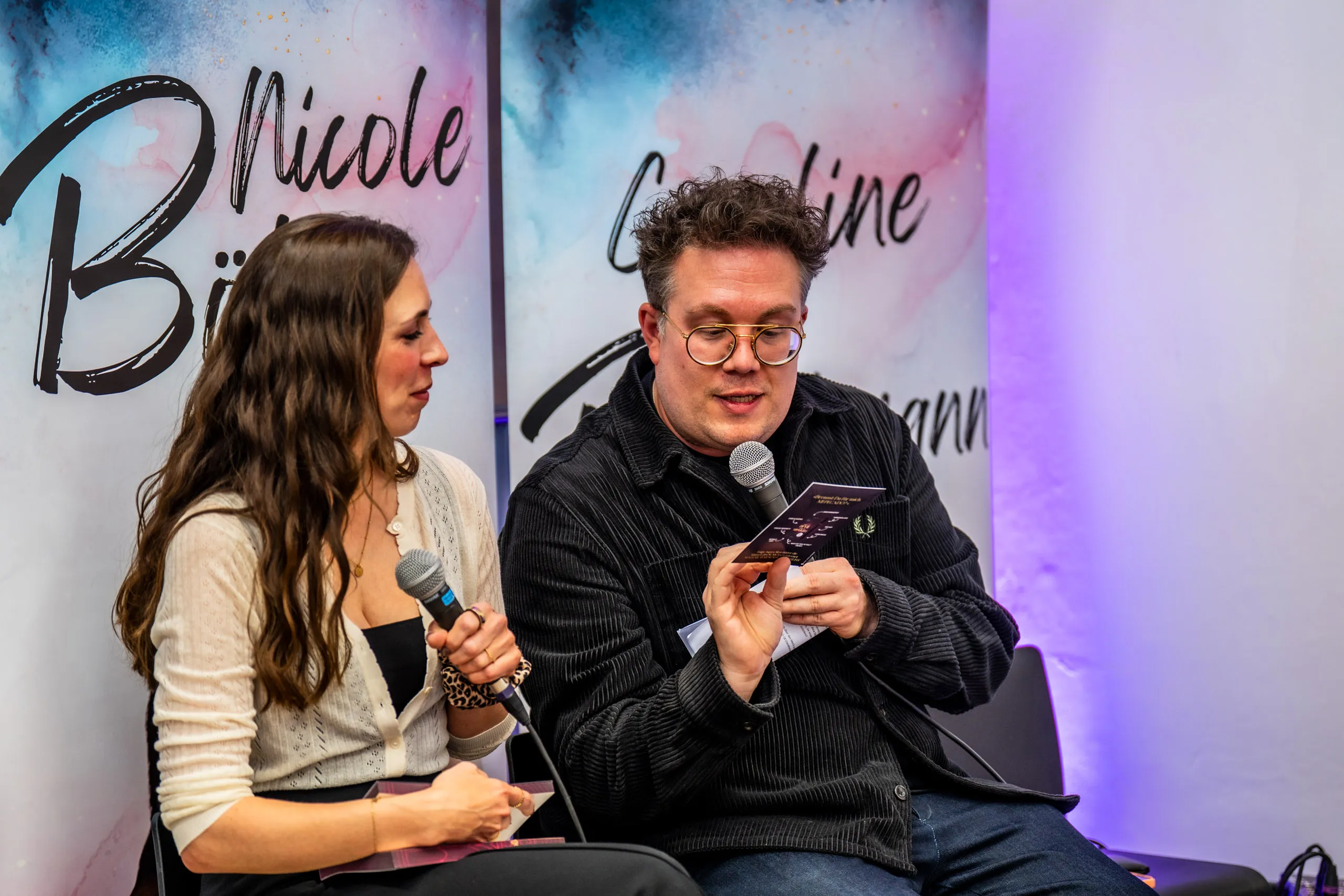 Thomas Sachsenmaier looking at a character card during the talk, Aspen Skye beside him with a microphone.