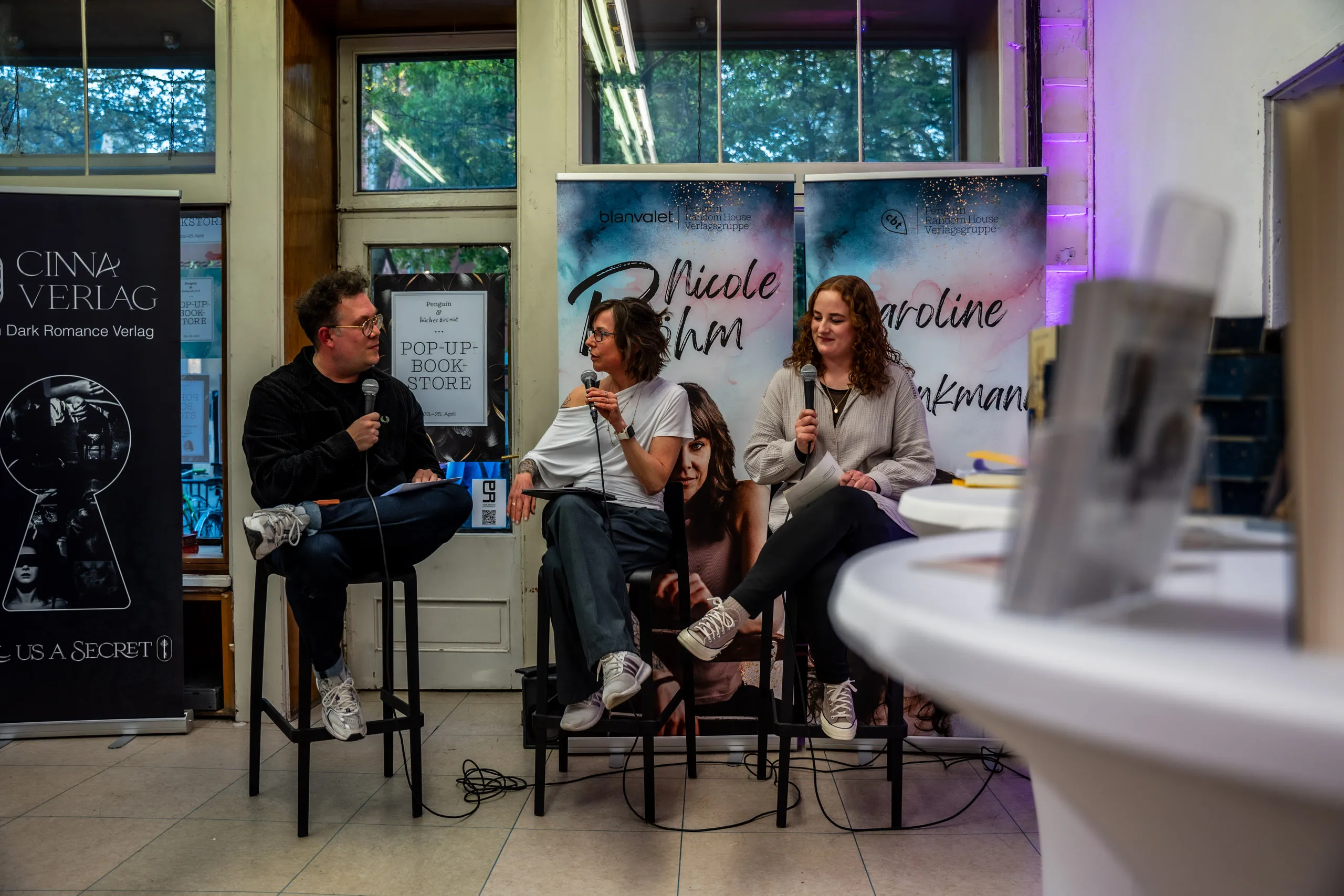 Talk at the pop-up book store: host Thomas Sachsenmaier in conversation with authors Nicole Böhm and Caroline Brinkmann, roll-up banners with their names and the CINNA Verlag behind them.