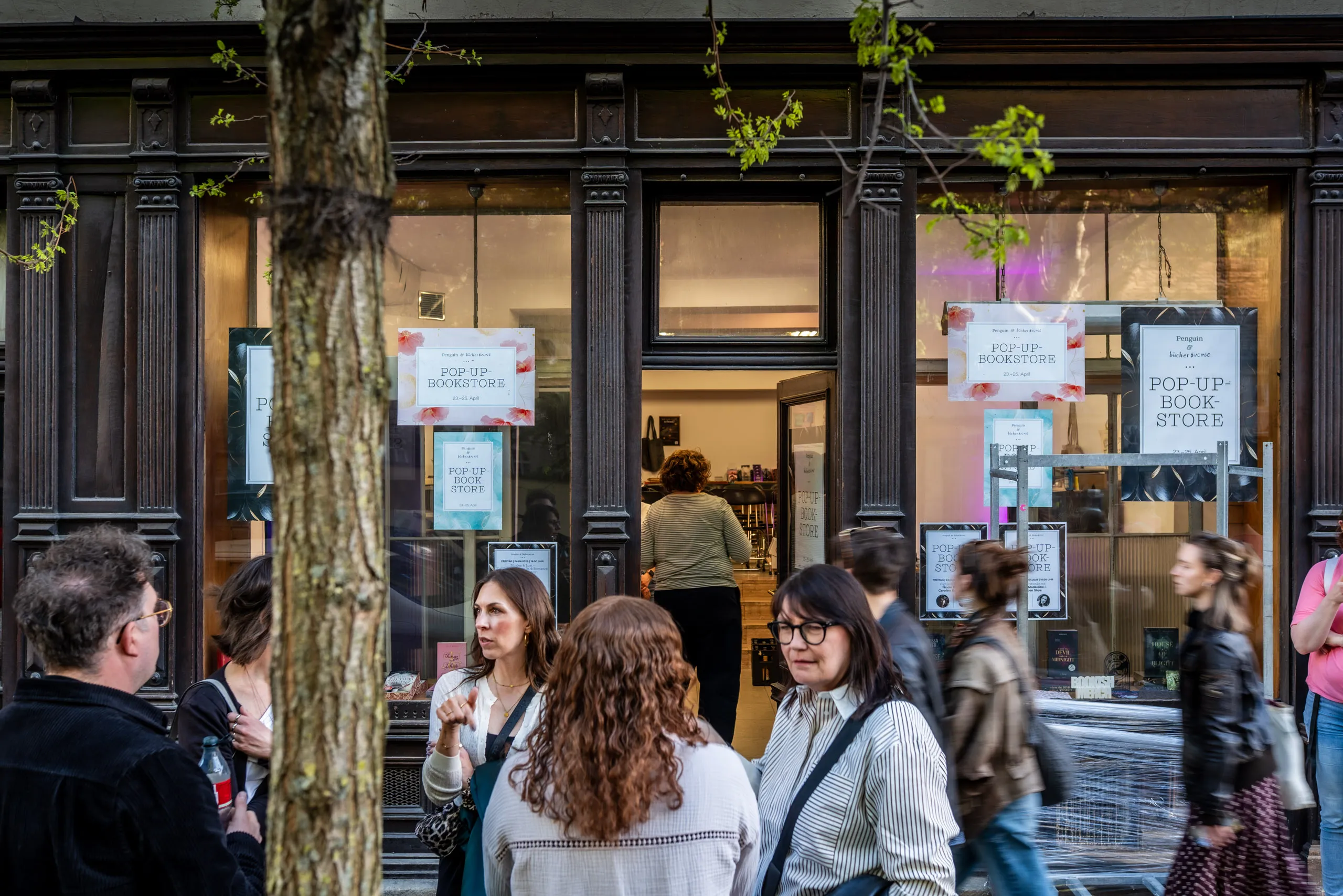 Besucherinnen und Besucher vor dem Penguin & Bücherbüchse Pop-Up Book Store in der Münchner Maxvorstadt, an der dunklen Holzfassade hängen Plakate mit der Aufschrift „Pop-Up-Bookstore".