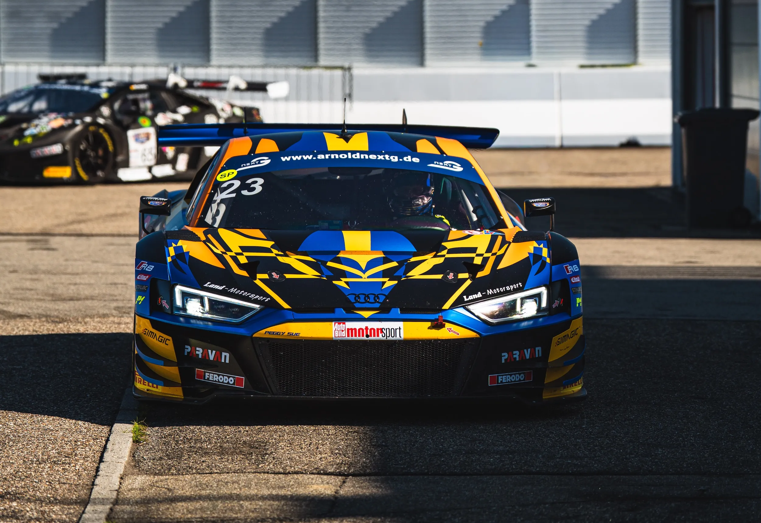 Der Audi R8 LMS GT3 von Land-Motorsport mit der Startnummer 23 rollt frontal auf die Kamera zu. Dahinter wartet ein dunkler Lamborghini Rennwagen vor der Tribüne des Hockenheimrings.