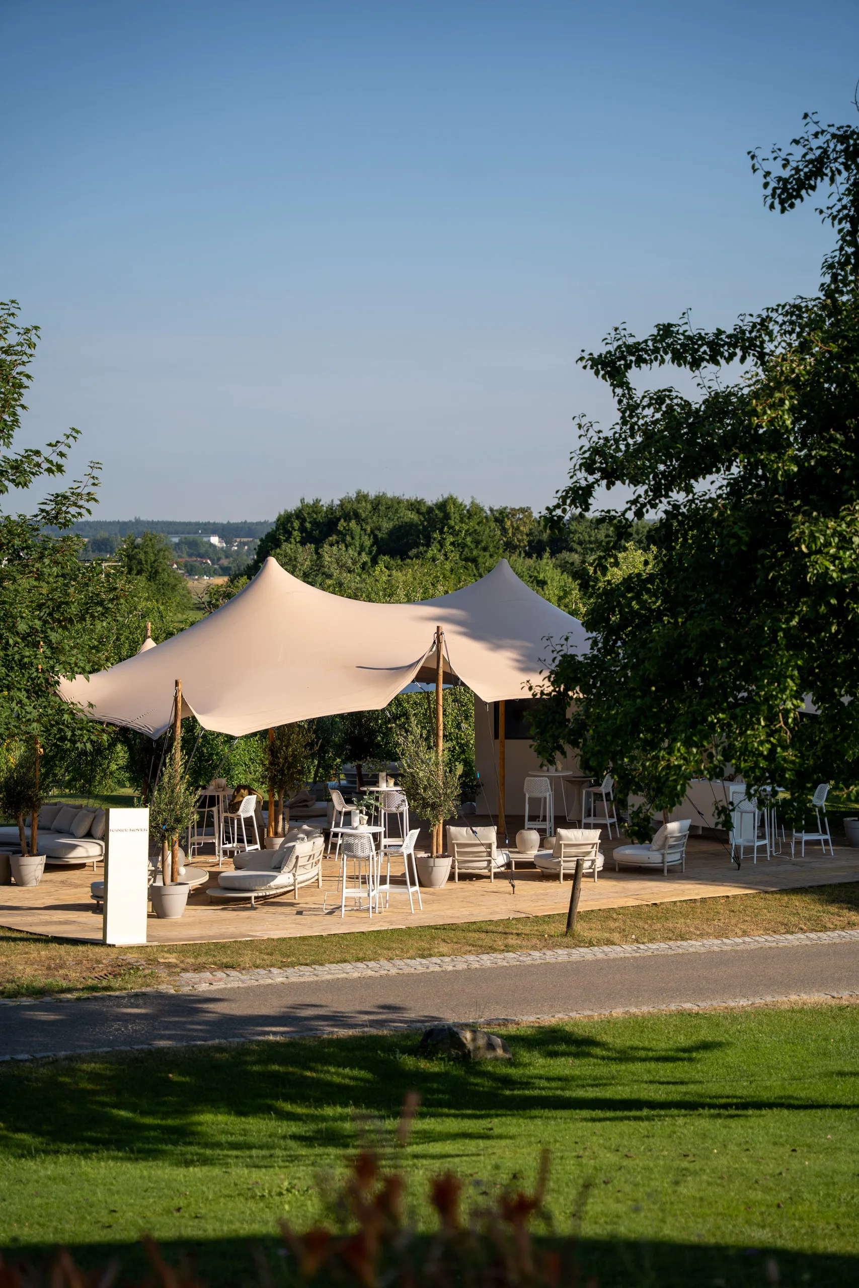 A stretch-tent lounge with white seating, standing tables and potted olive trees on a wooden deck; a Range Rover plinth stands at the left, with meadows and trees beyond.