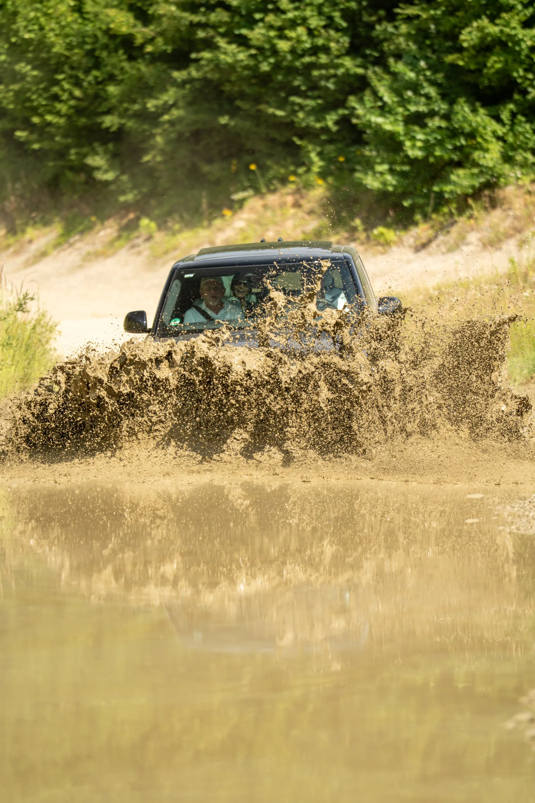 A Range Rover Sport ploughs through a mud hole, throwing up a wide brown spray. Several occupants are visible through the windscreen.