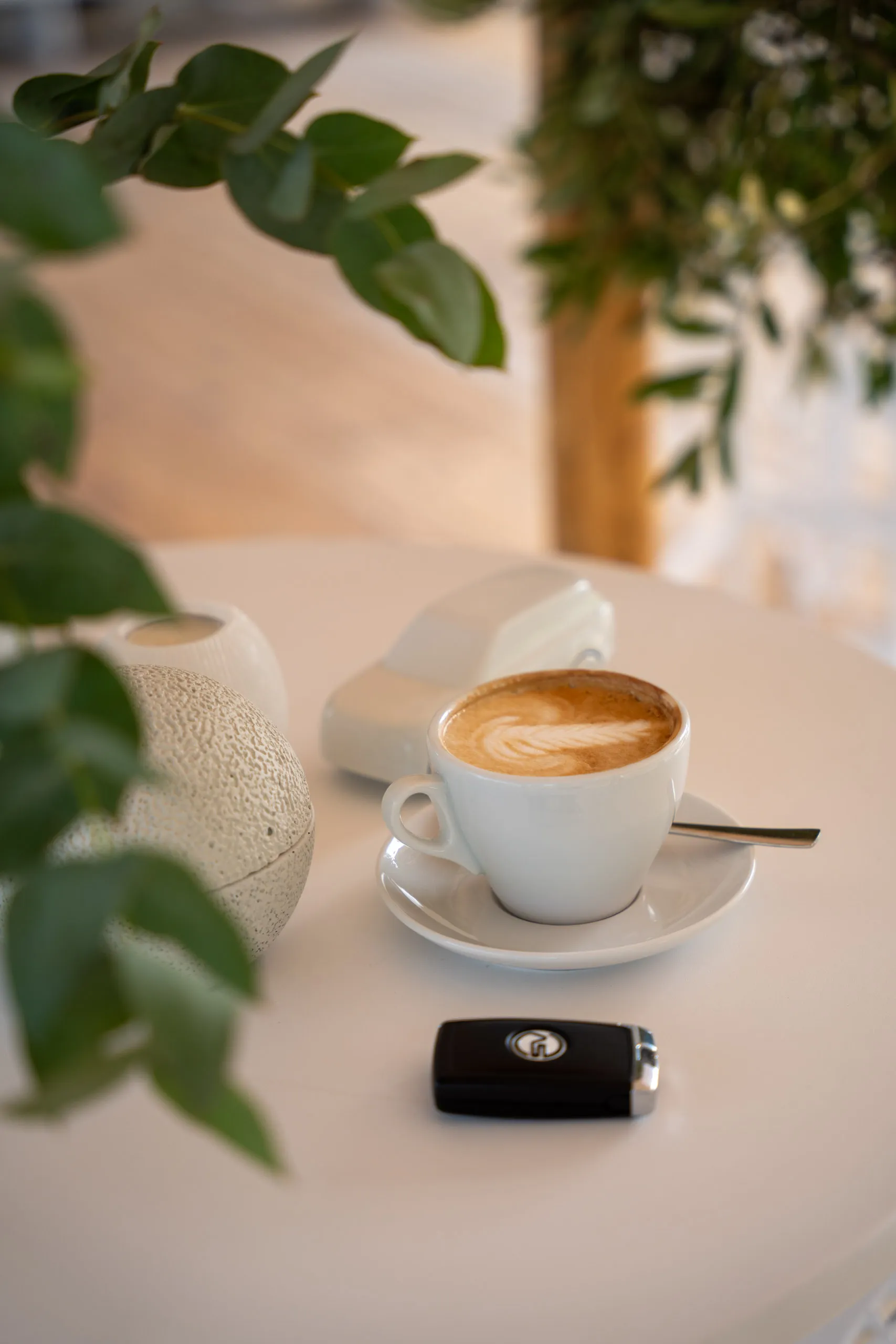 A cappuccino with latte art sits on a pale table, a Range Rover SV key in front of it and a white model car behind, framed by eucalyptus branches.