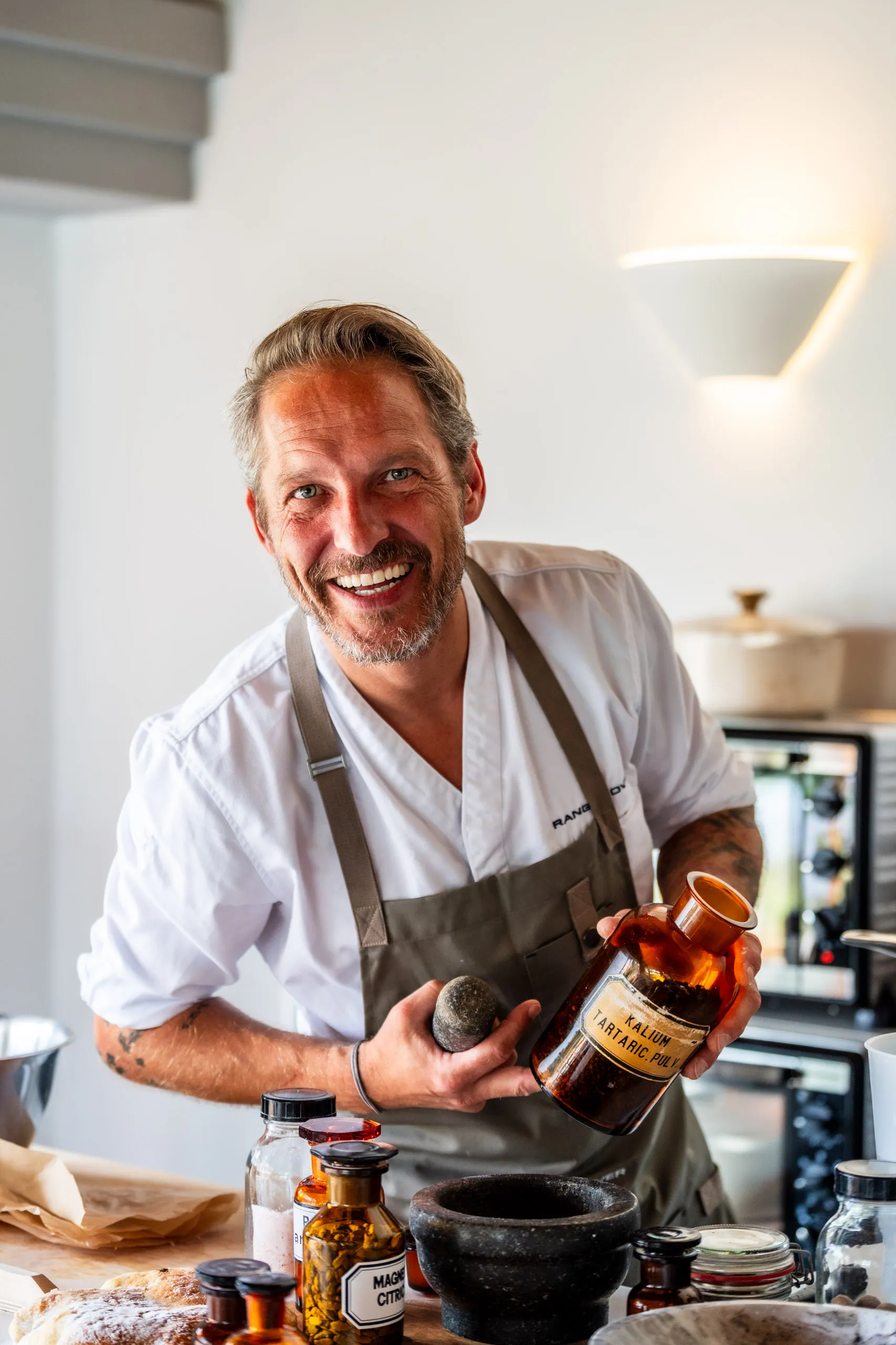 Rainer Klutsch, in a white chef’s jacket embroidered with Range Rover, laughs as he holds a brown apothecary bottle labelled "Kalium tartaric. pulv." and a stone pestle, a mortar and labelled jars in front of him.