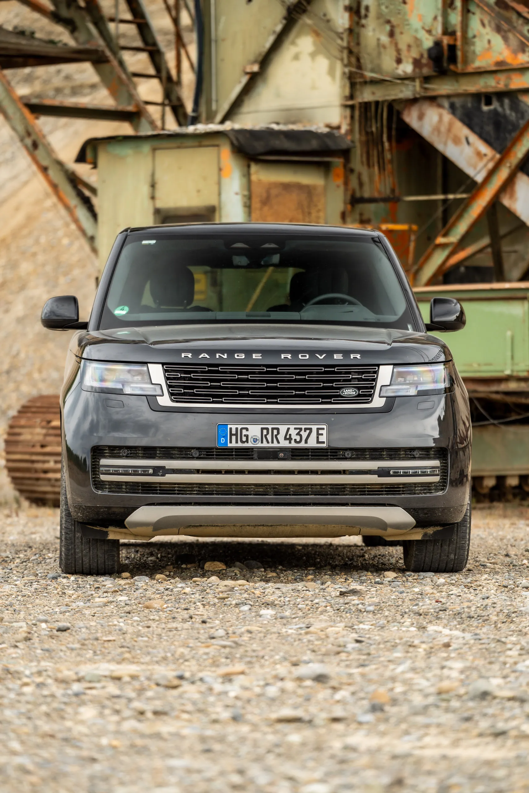 A Range Rover stands head-on on gravel, with the rusting steel structures and conveyors of a gravel pit behind it.