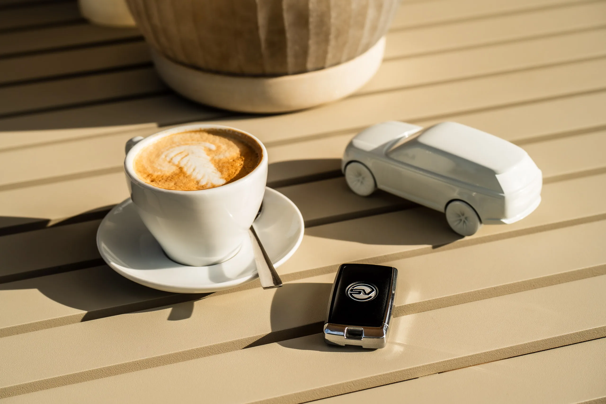 A cappuccino, a Range Rover SV key and a white model car sit side by side on a beige slatted table in warm sunlight.